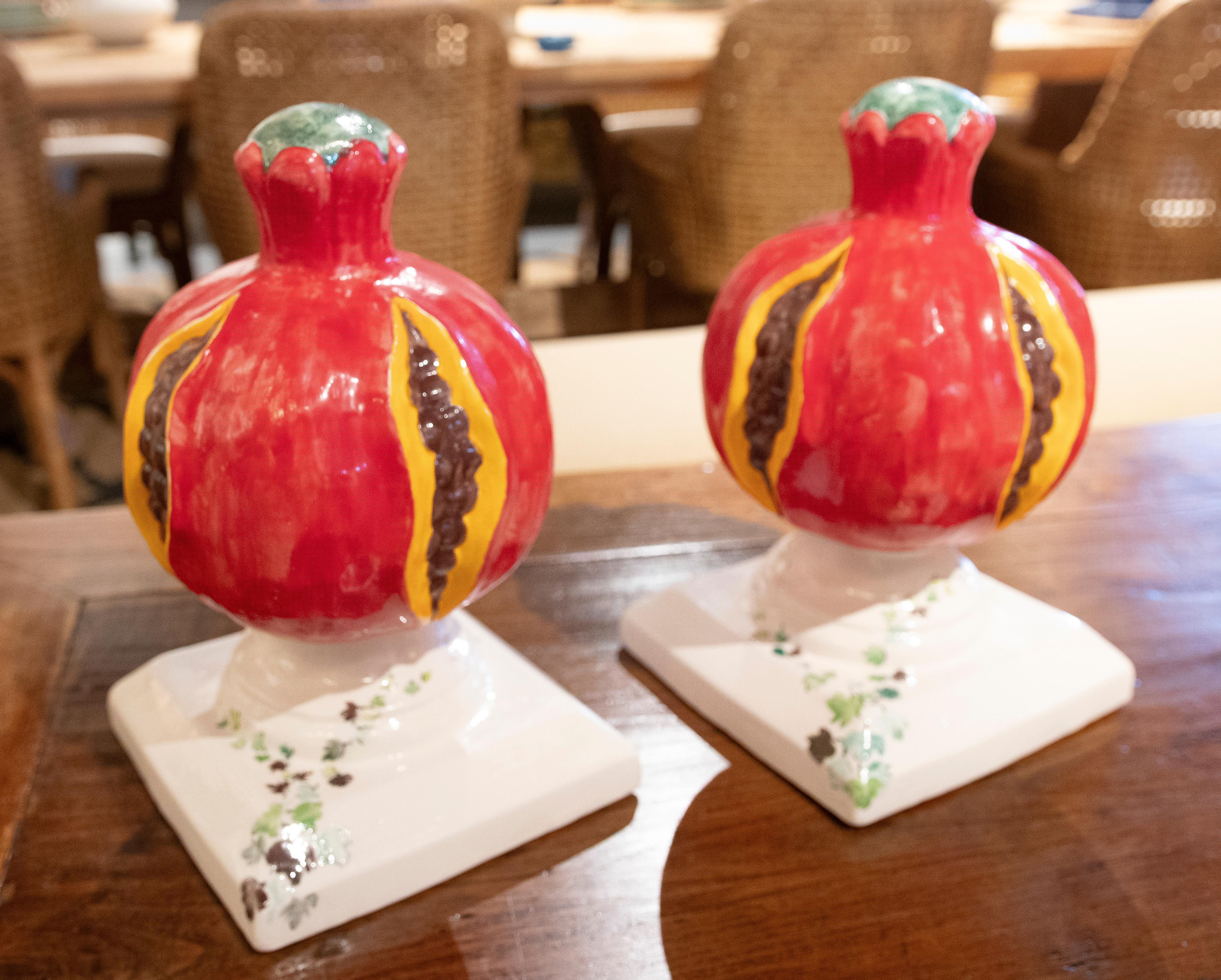 A charming pair of glazed ceramic decorative sculptures depicting open pomegranates with embossed details and vivid colors. The fruits, glazed in glossy red with yellow accents and brown seeds, rest on elegant square bases with hand-painted floral