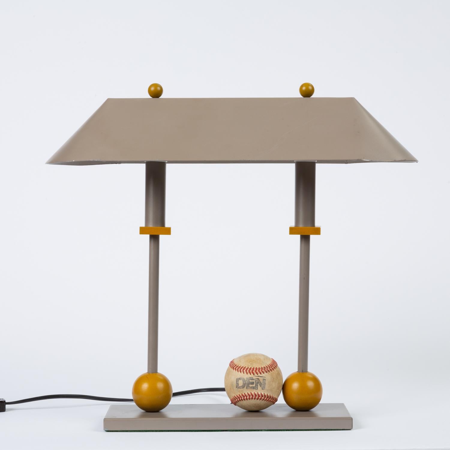 A Postmodern iteration of the Classic two-pillar banker’s lamp from lighting designer Robert Sonneman for George Kovacs. Designed in 1990, this piece has a triangular shade in putty-gray enameled aluminum, supported by two columns on a flat base.