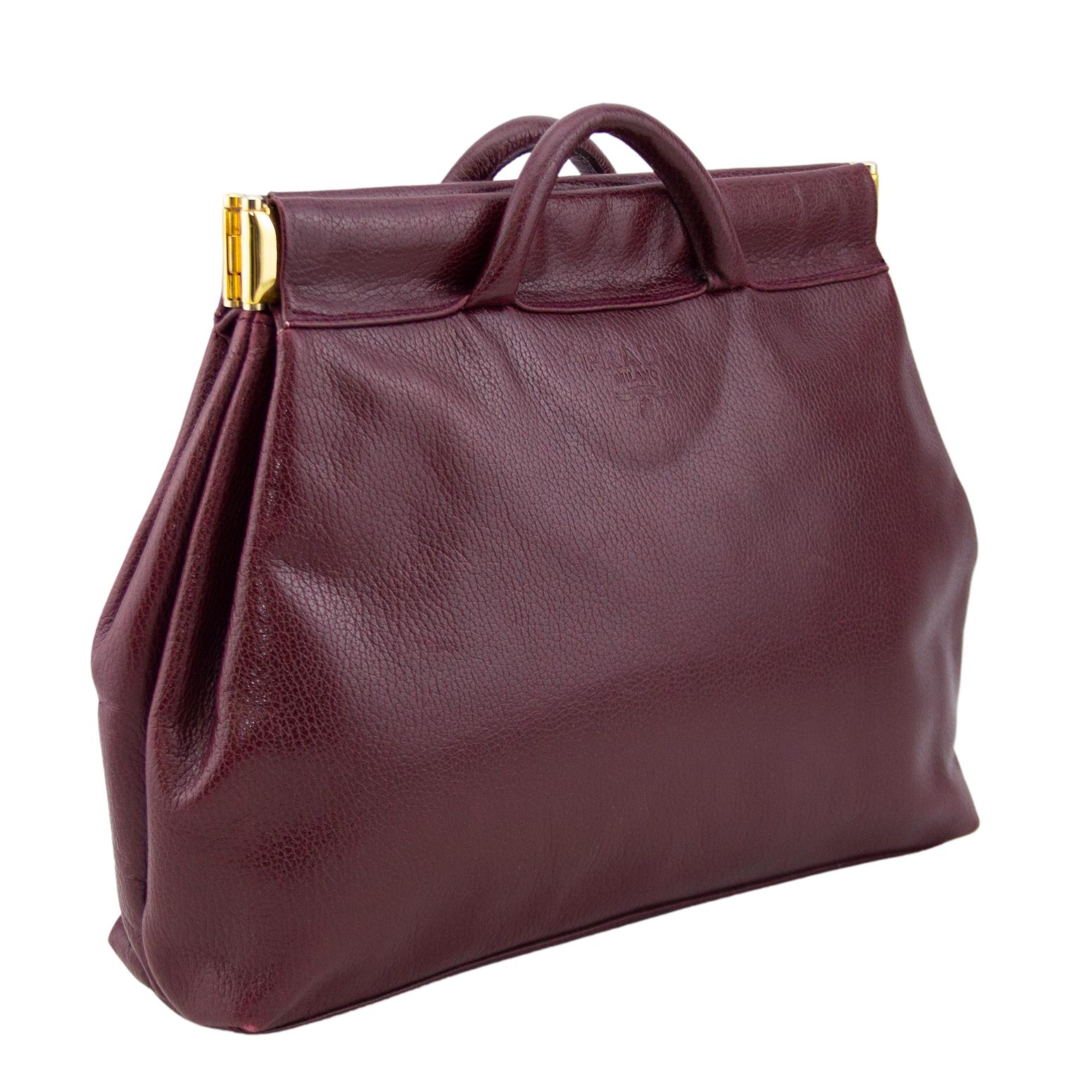 Early 1990s Prada clutch. Maroon rigid leather with gold hardware and very small handles. Bag is made up of 2 pockets and stiff interior hardware keeps bag sturdy and open when expanded and snaps shut. Exterior Prada Milano blind stamp in leather.