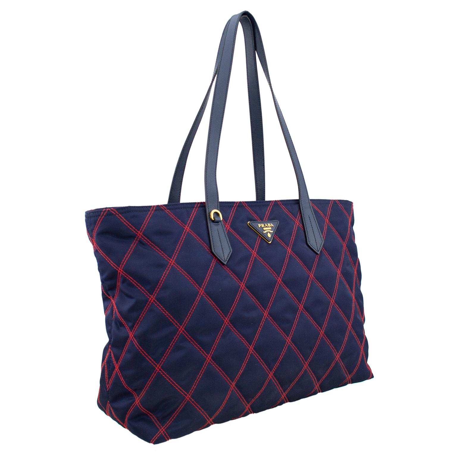 Prada tote bag from the 1990s. Navy blue nylon with red quilted top stitching and triangular Prada logo. Blue leather flat handles that sit comfortably on the shoulder. Prada logo lined interior with two open and one zippered slit pocket. Zipper