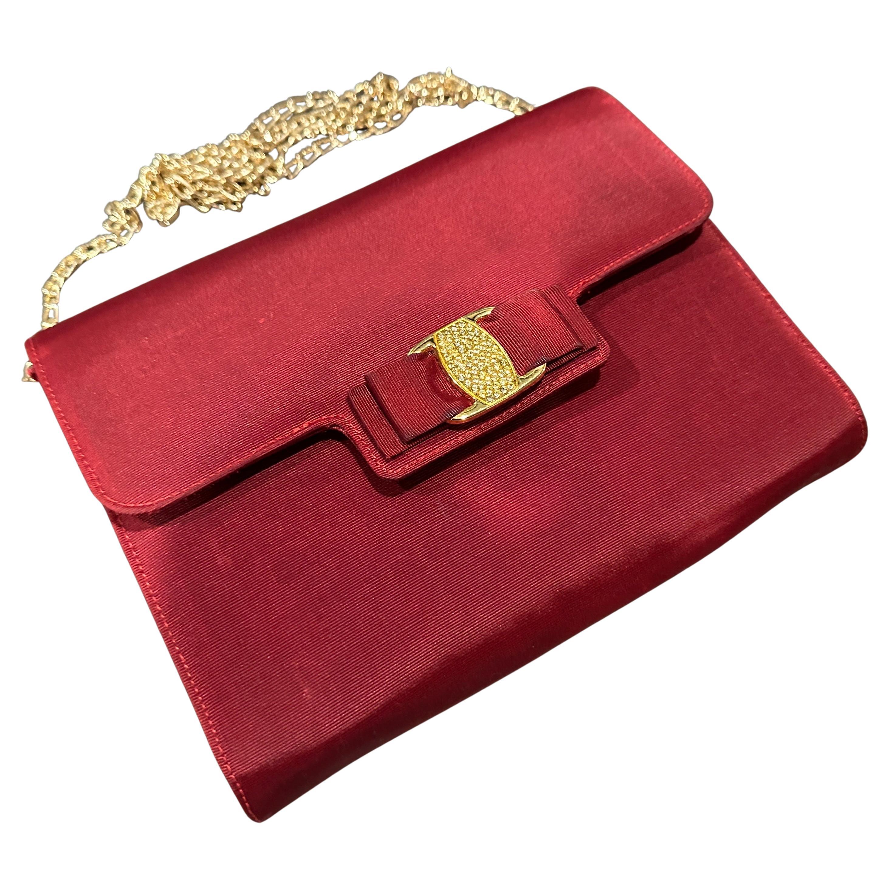 This is a vintage evening bag from the 1990s by Salvatore Ferragamo, crafted in a rich red silk that gives it a smooth and elegant texture. The design is structured and timeless, featuring a signature Vara bow on the front with a gold-tone brass