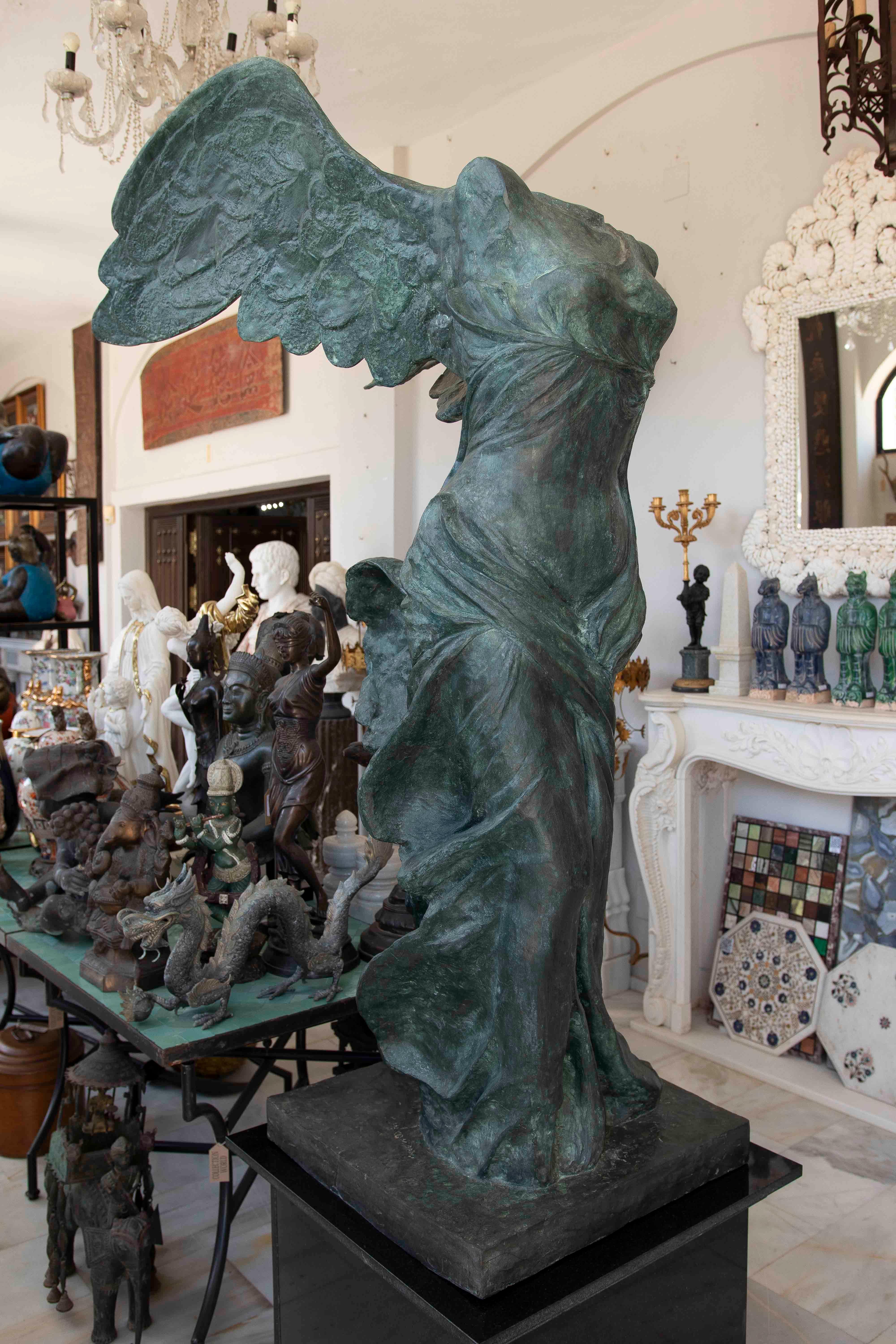 An imposing reproduction of the famous Victory of Samothrace, cast in bronze with a green patina finish that highlights the folds and energy of the figure's movement. The sculpture depicts the iconic winged pose, with the torso tilted and the