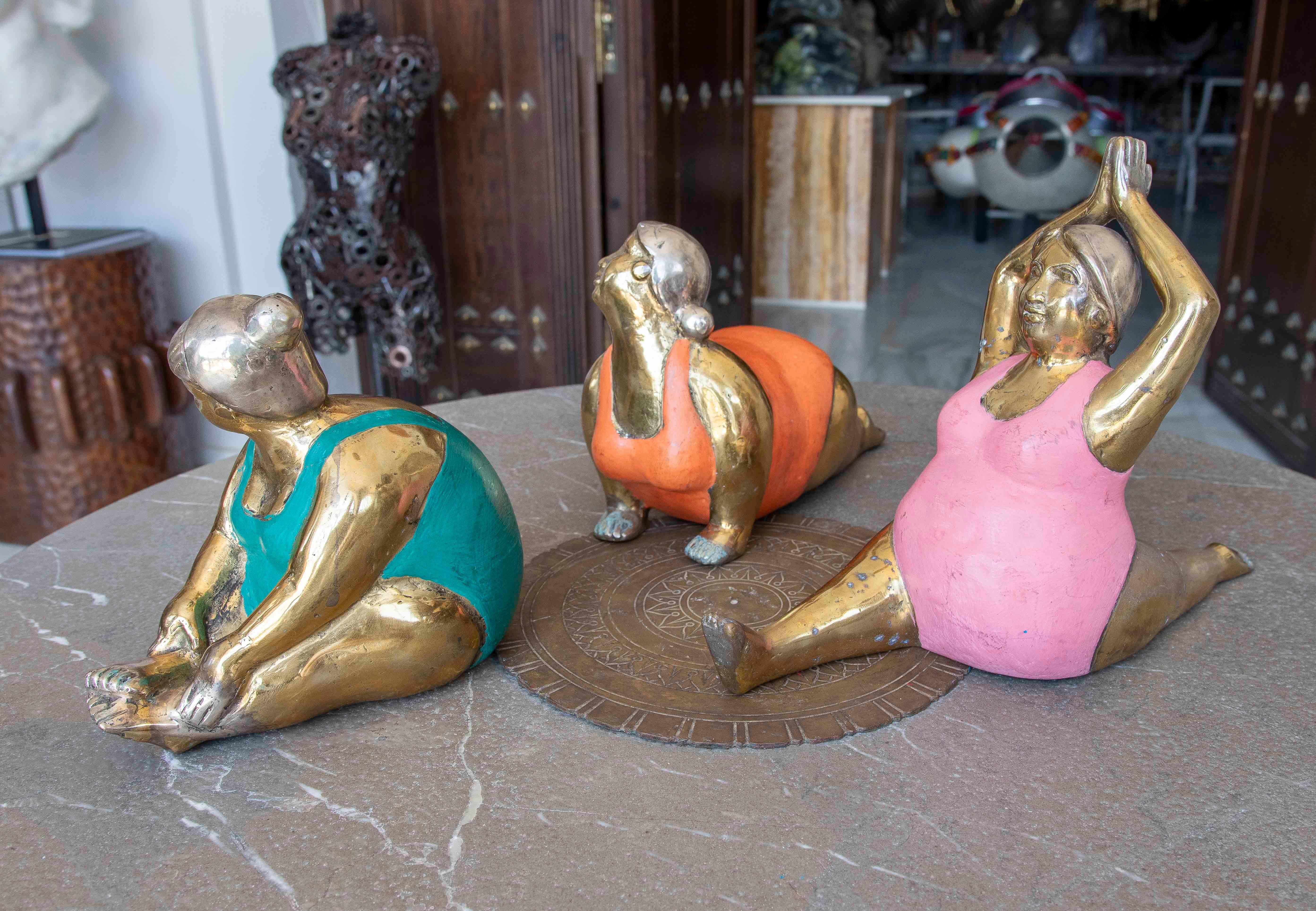 A set of three bronze sculptures depicting robustly built women performing various yoga poses. Each figure wears a brightly colored swimsuit (green, orange, and pink), elegantly standing out against the body's shiny gold finish. These pieces combine
