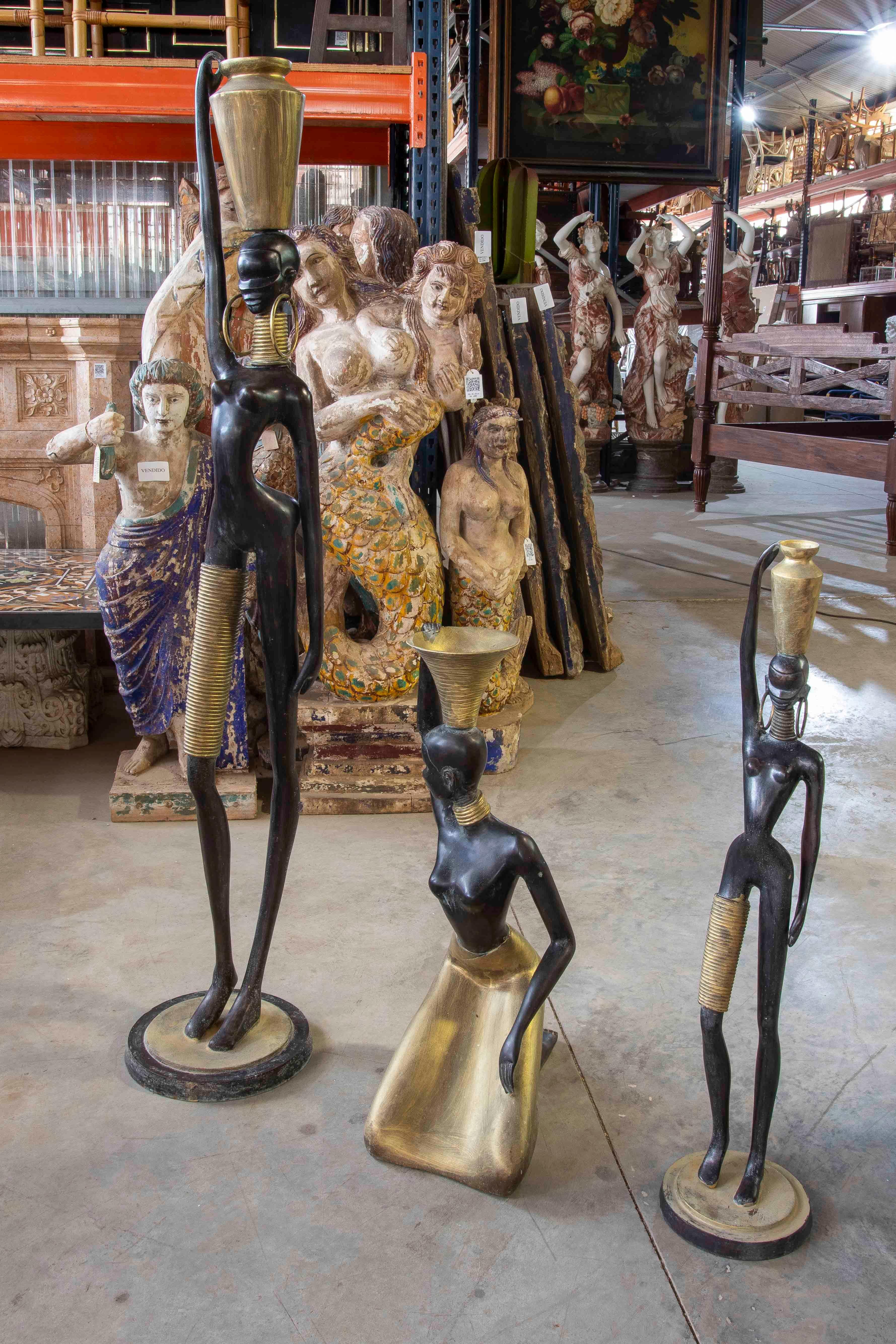 Conjunto decorativo de tres esculturas africanas modernas de bronce patinado en negro con detalles dorados. Las figuras representan a mujeres estilizadas que portan jarras y llevan adornos tradicionales como collares y faldas metálicas. El diseño