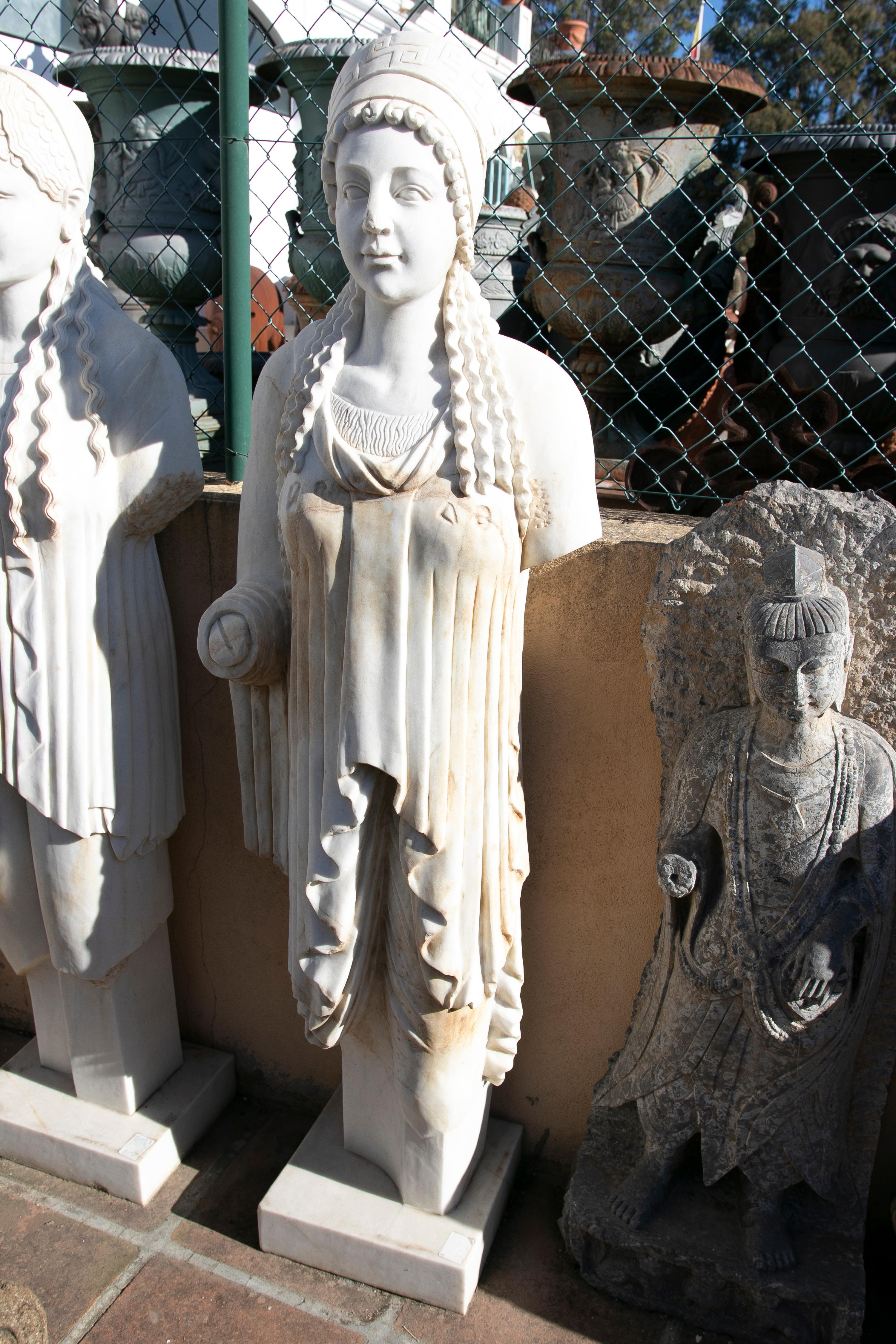1990s hand carved aged Macael white marble reproduction of Classical Greek Kore female sculpture. Its posture is rigid and column-like, showing the restrained 