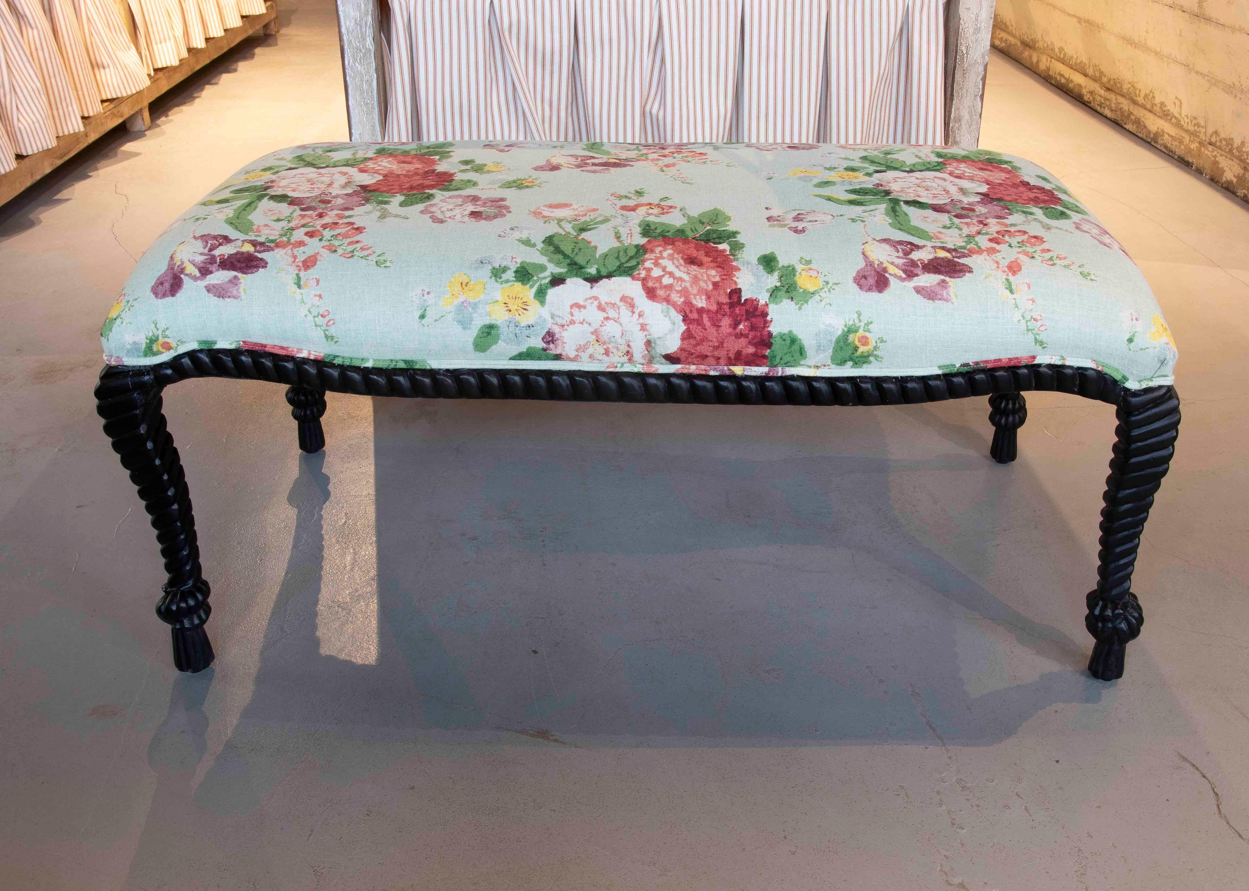 Banc vintage élégant avec un cadre en bois et des pieds tournés noirs, lui conférant caractère et sophistication. L'assise est recouverte d'un tissu floral dans des tons pastel, principalement des verts tendres, des roses, des jaunes et des touches