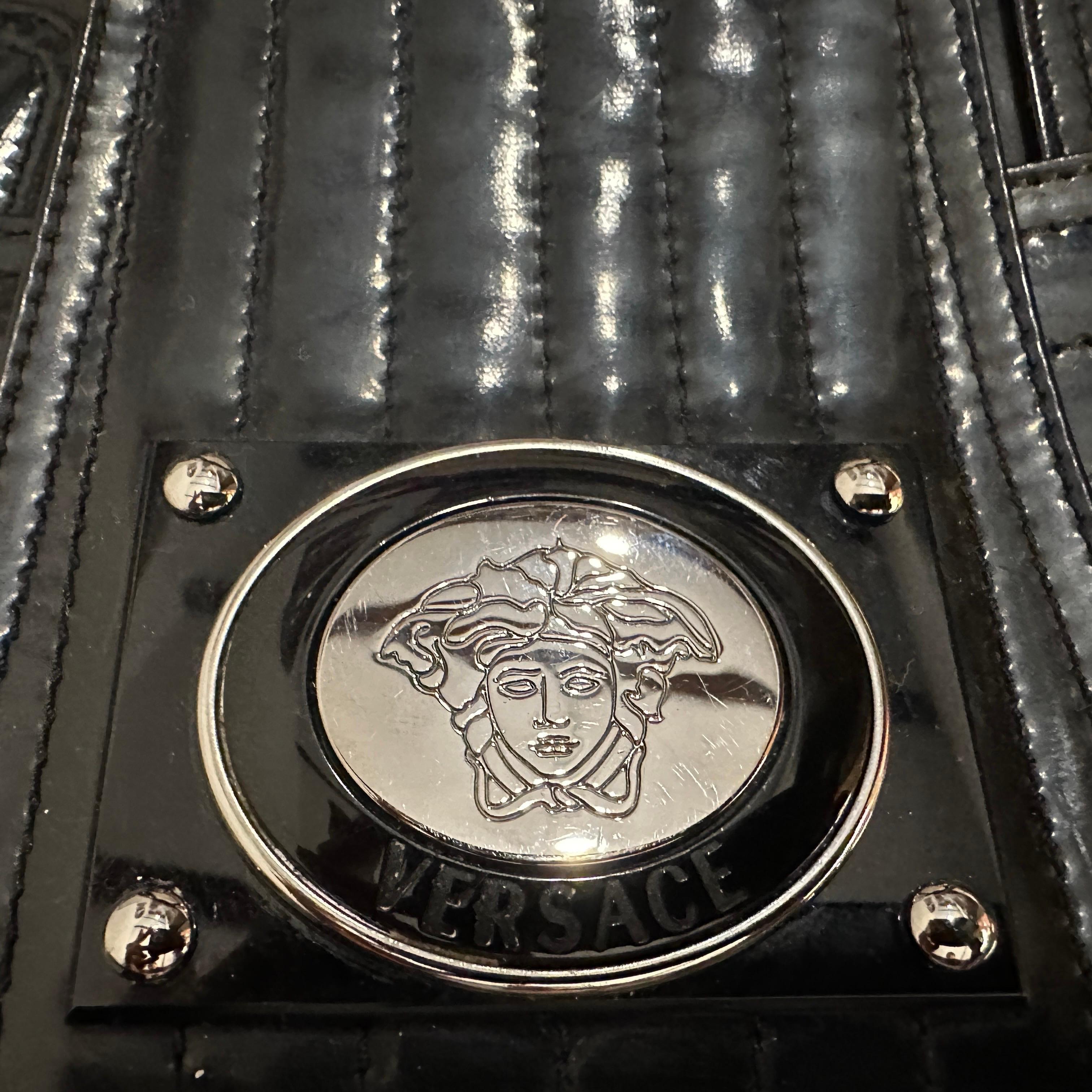Ce sac à main vintage des années 1990 de Gianni Versace reflète le style audacieux et extravagant qui définissait la marque à cette époque. Le sac est fabriqué à partir d'une combinaison de cuir verni noir brillant et de plastique, ce qui lui