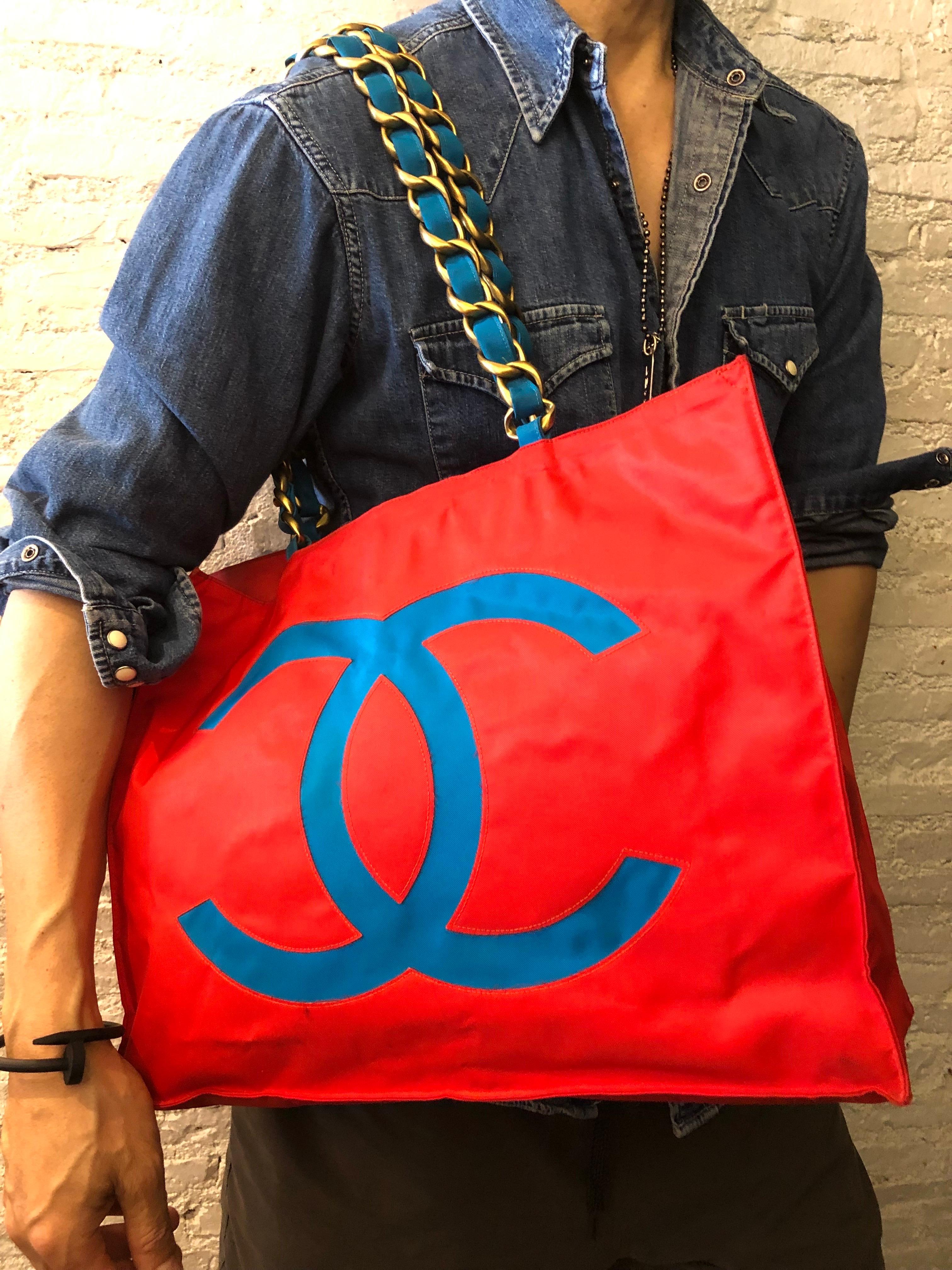 This vintage CHANEL jumbo chain tote is crafted of nylon in red and turquoise featuring a massive cut-out CC logo on one side and cut-out CHANEL letters on the other in turquoise. This tote has two sturdy gold toned shoulder chains interlaced with