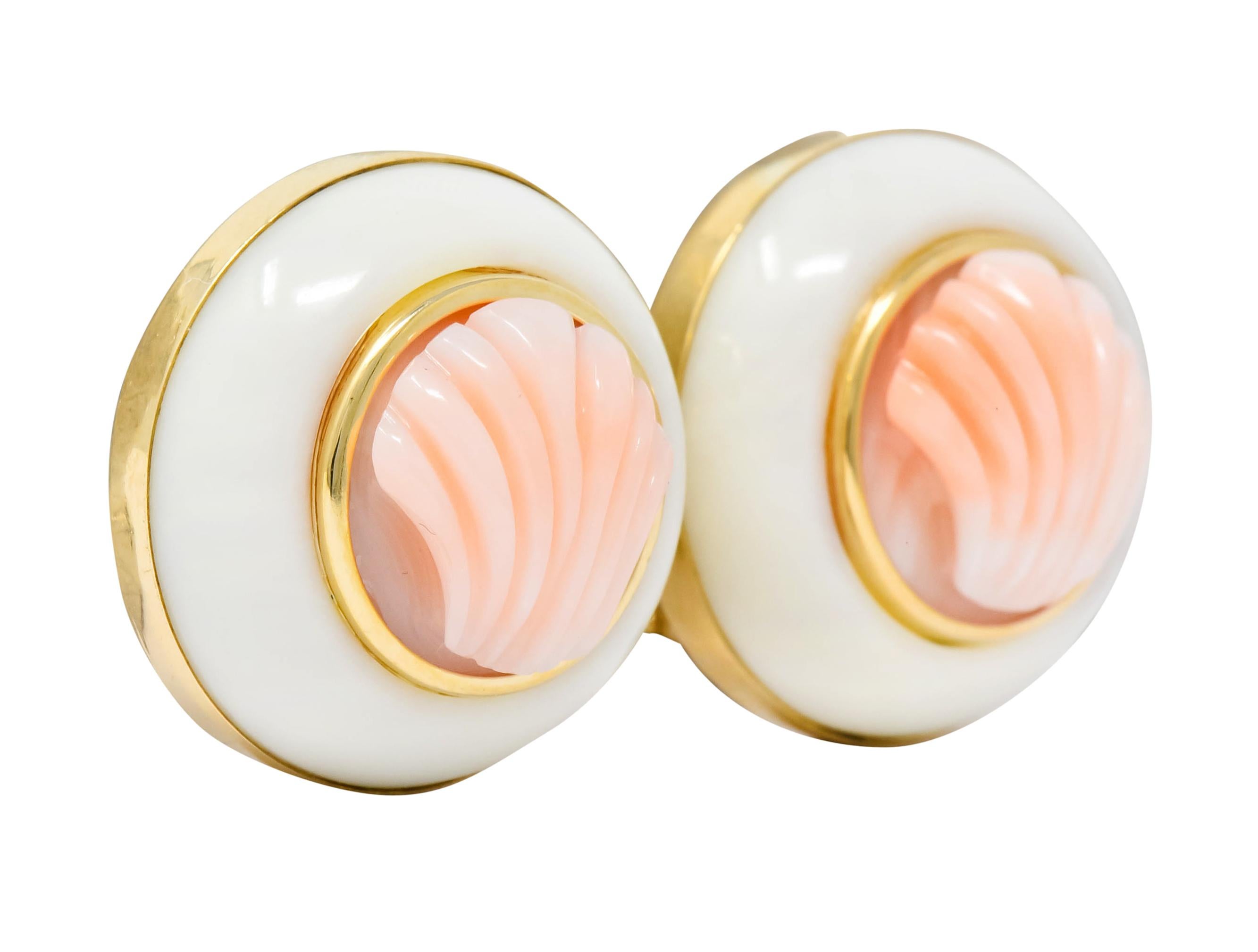 Each earring centers deeply carved coral, very light peachy-pink, depicting a fanned shell motif

With a second circular ring of coral cabochon, opaque and creamy white in color

Both bezel set in polished gold surrounds

Completed by posts and