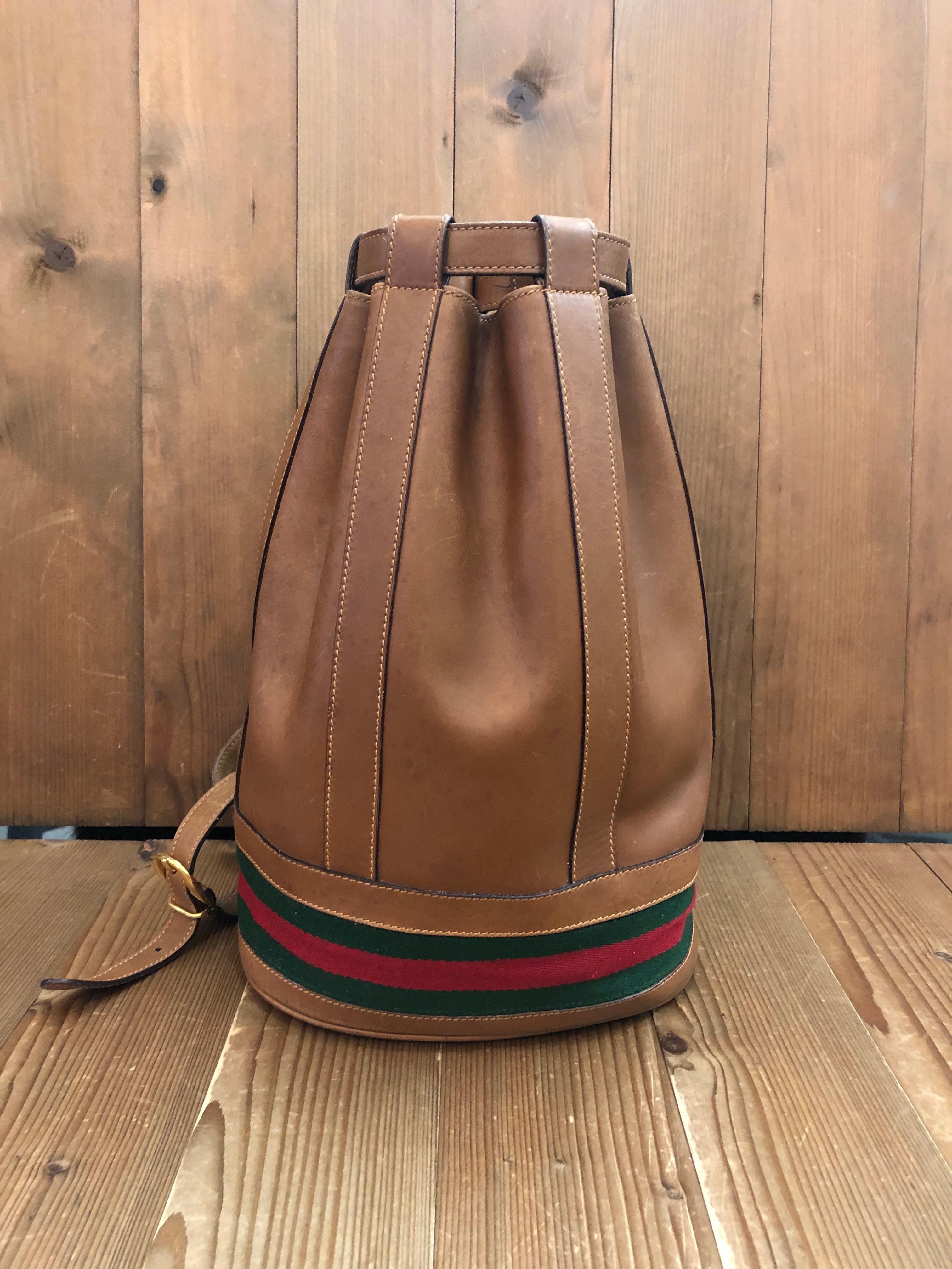 This vintage GUCCI Web bucket shoulder bag is crafted of smooth calfskin leather in tan featuring gold toned hardware and decorated with Gucci’s iconic red/green stripe around the bottom. Top drawstring closure opens to a new coated interior in