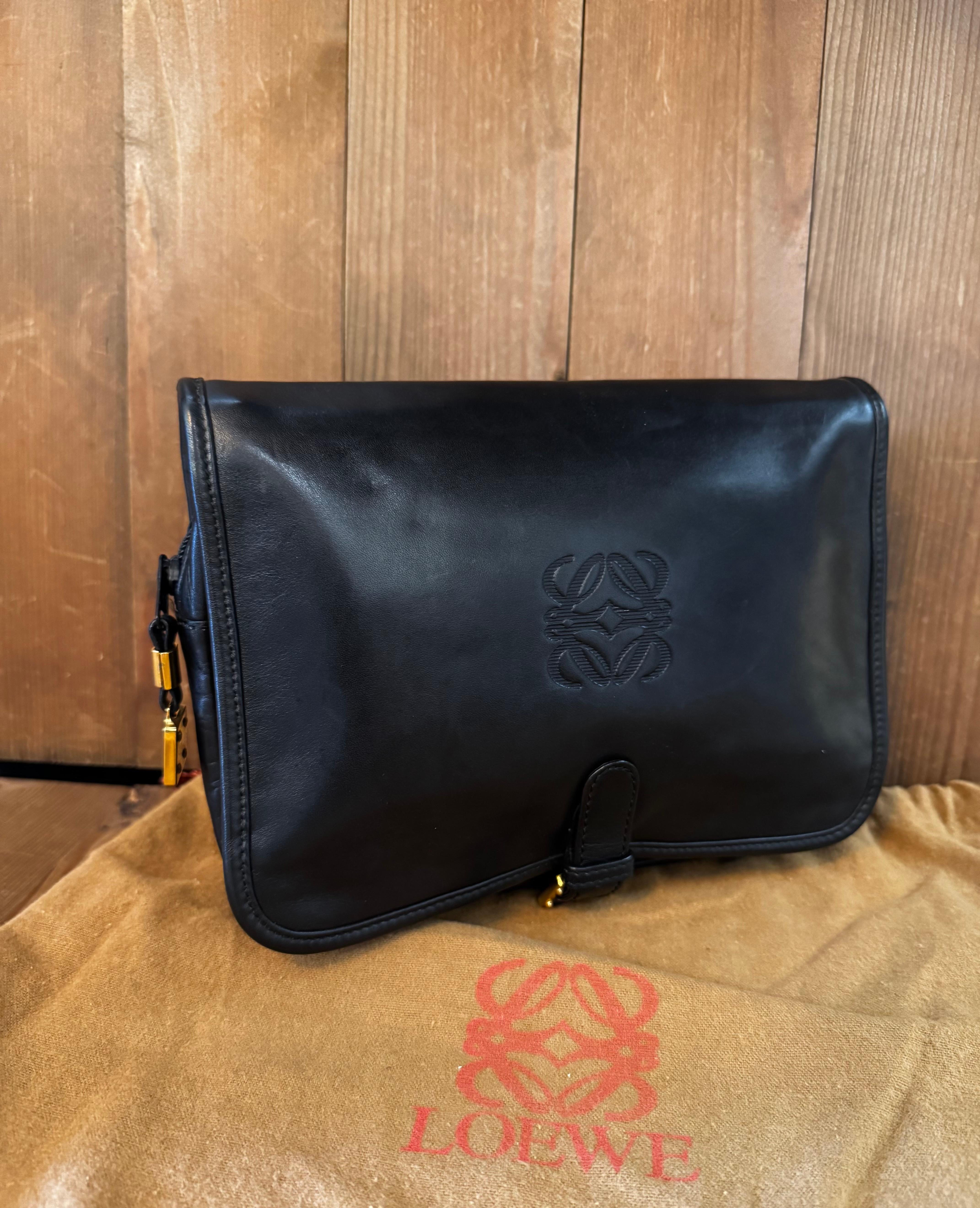 Cette pochette vintage à deux volets de LOEWE est confectionnée en cuir de veau souple et lisse, de couleur noire et ornée de ferrures dorées. La fermeture à boucle inférieure permet d'accéder à deux poches zippées et à une poche principale zippée.