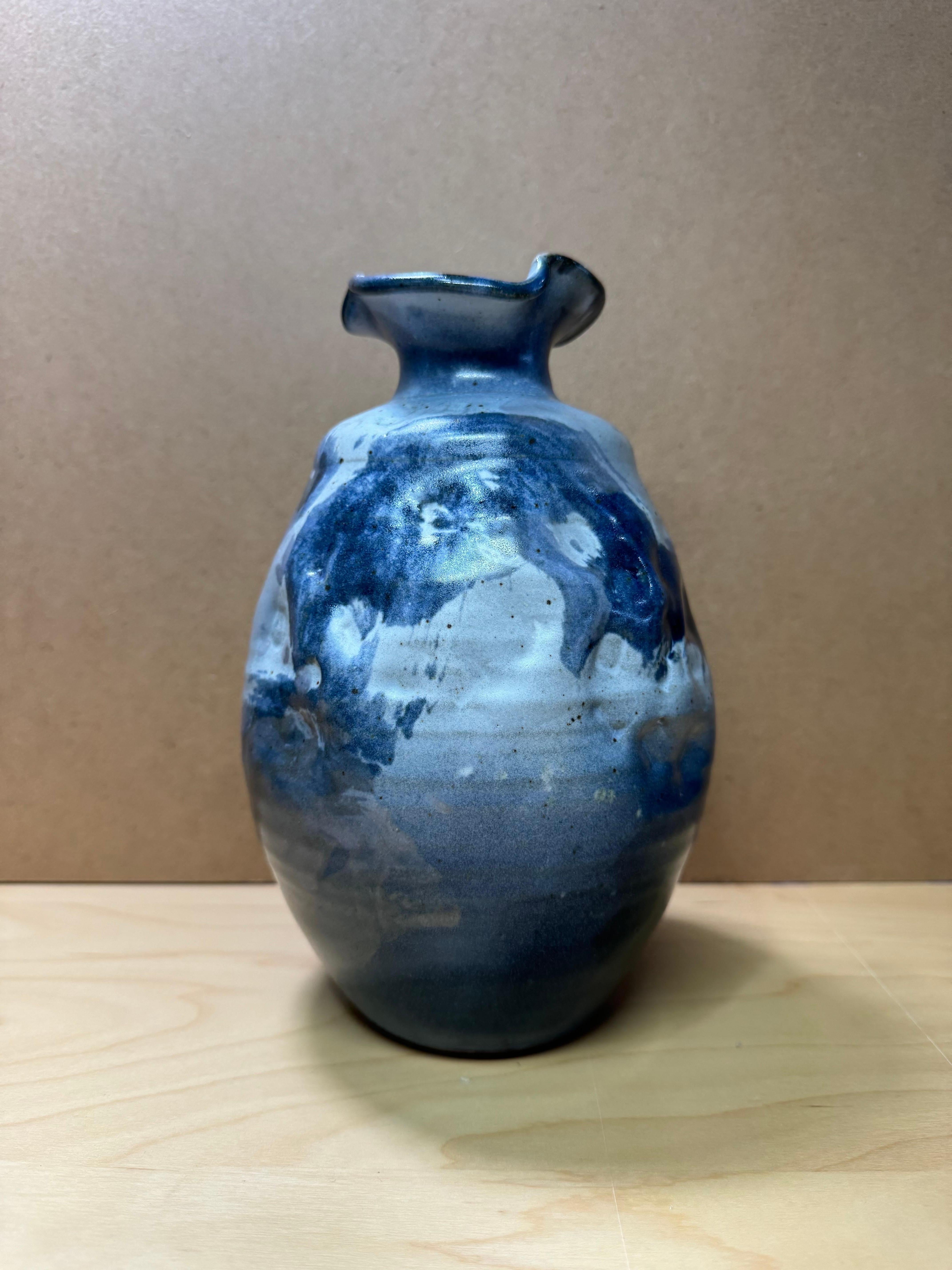 Beautiful hand thrown pottery vase with fluted and ruffled rim and uneven body. Finished with different shades of blue, this vase is a stunner and eye-catcher that will draw attention wherever displayed.
This vase is signed 