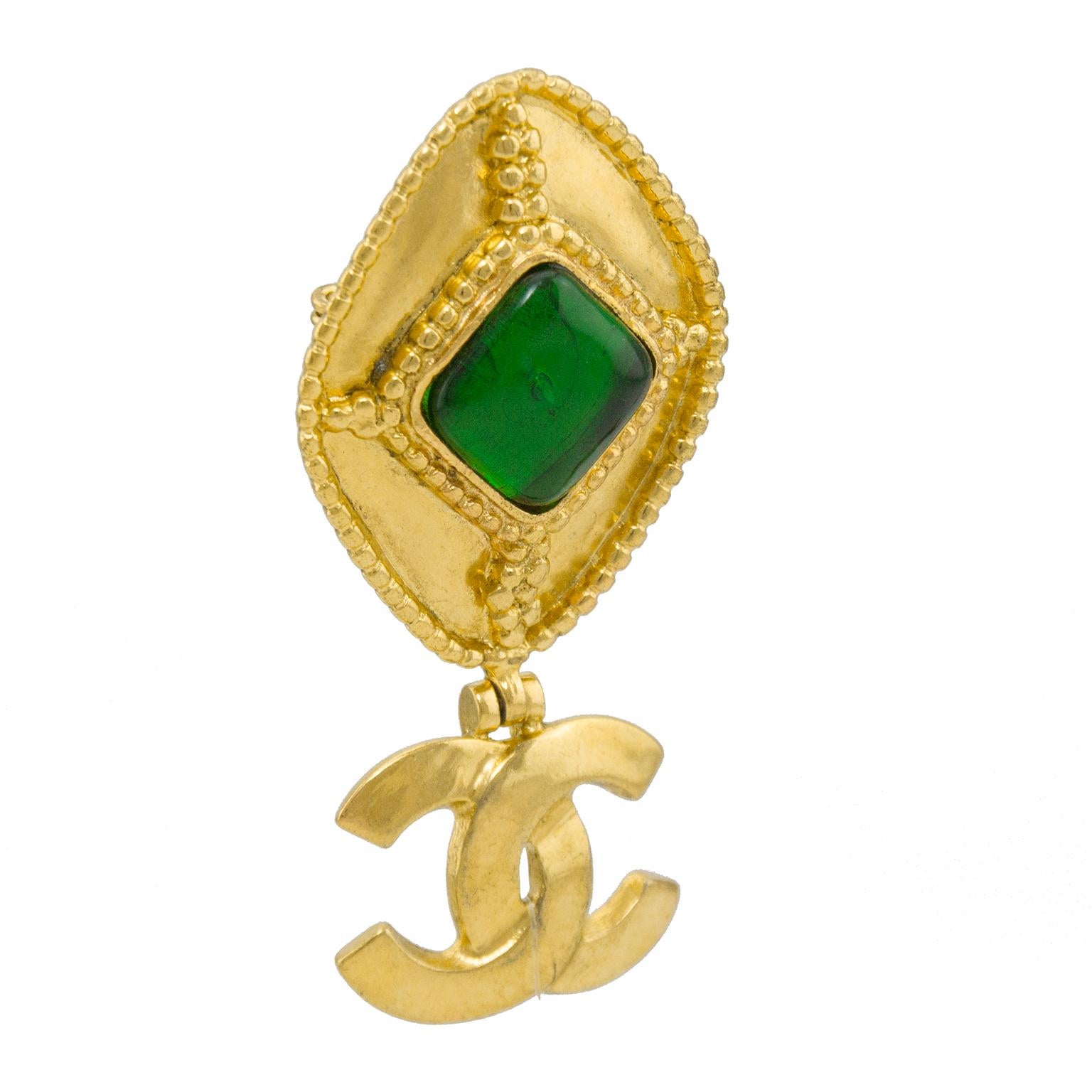 Chanel pin from the Fall 1996 collection. The diamond shaped pin has a green poured glass center stone surrounded with an intricate bead border. A Chanel CC charms drops off the bottom point and has a hinge attachment allowing the CC to move along