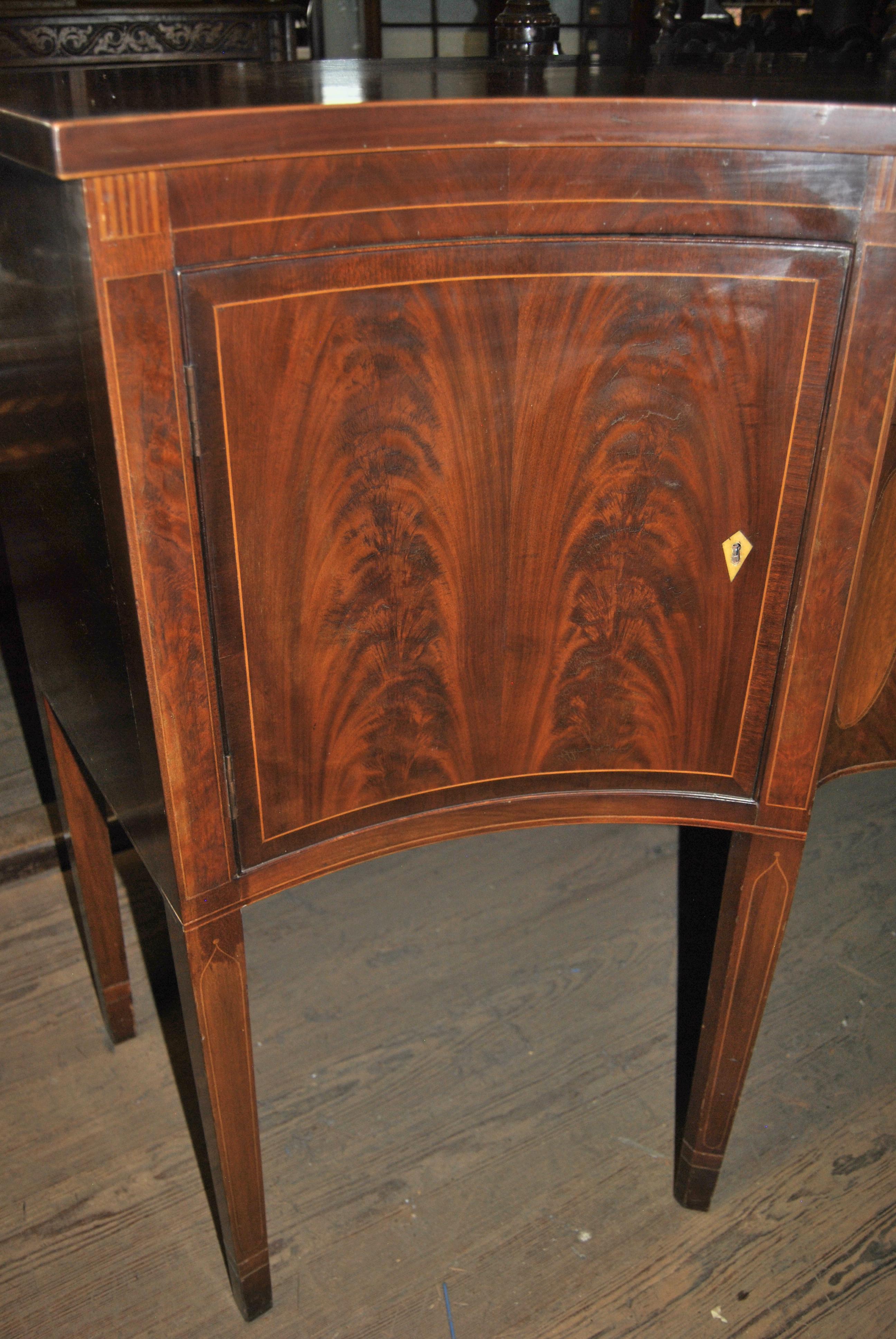 This is a mahogany sideboard, server, buffet made in England. The top is shaped to fit the form of the base and is concaved on the right and left sides with a bow front to the middle. The edge of the top has a String Inlay of Satinwood. Each of the