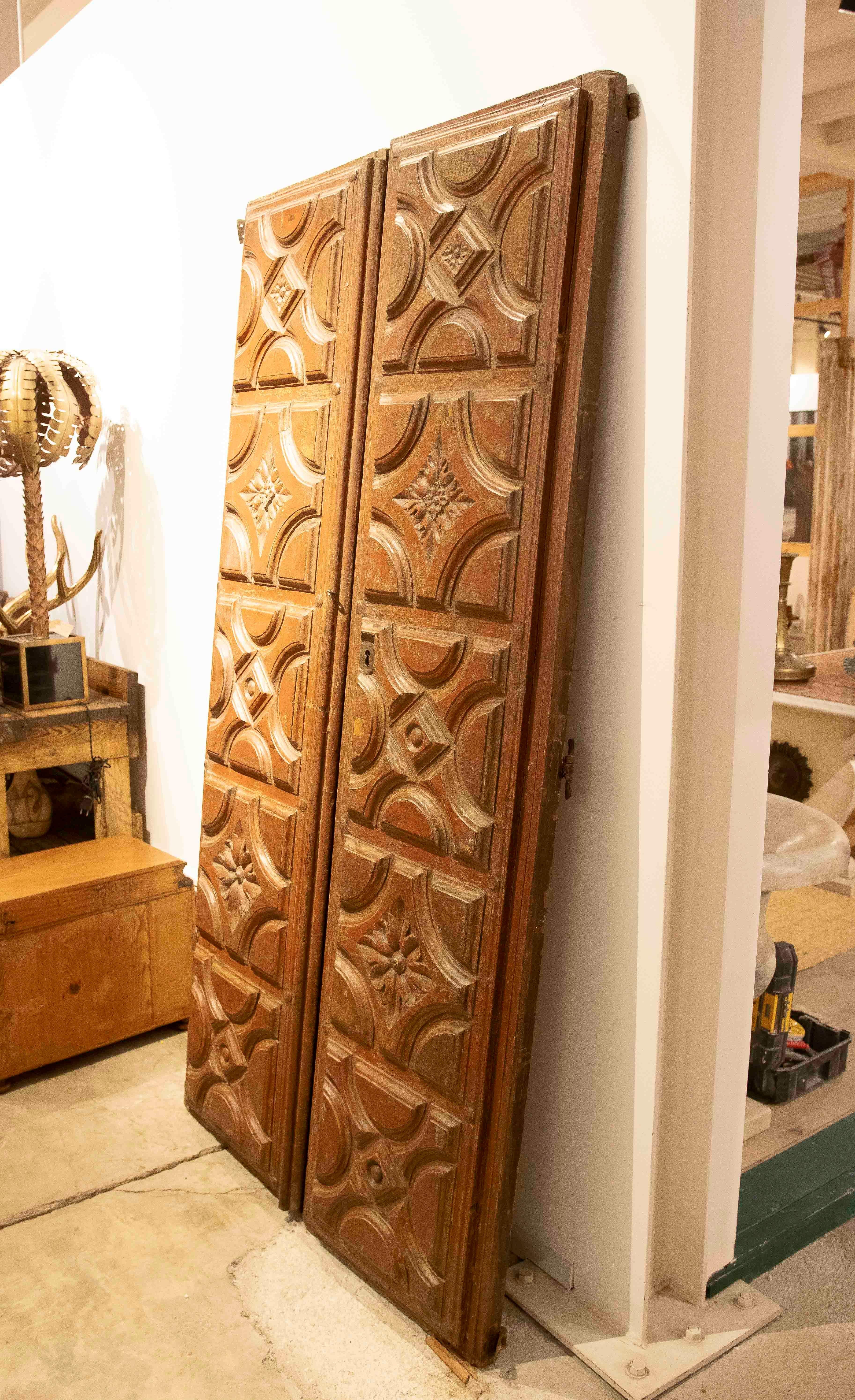 Solid wood double-leaf door, decorated with relief-carved panels alternating semicircular and rhomboidal geometric shapes, each centered by a hand-sculpted floral motif. The symmetrical arrangement of the elements, along with the slightly worn