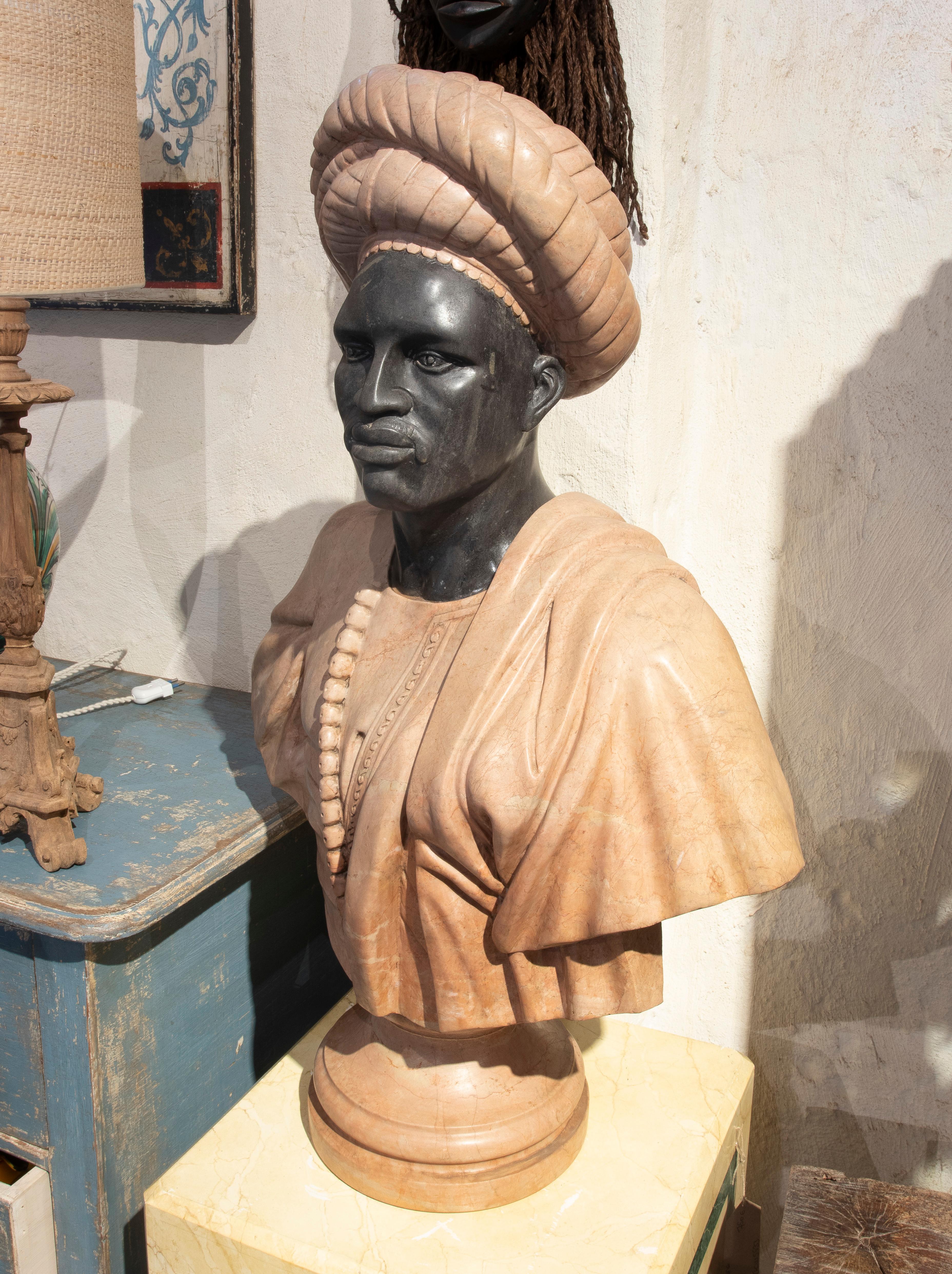 Magnificent hand-carved marble bust depicting an African gentleman in traditional attire and a detailed headdress. The work stands out for its use of two-tone marble, with a contrast between the polished dark marble face and the pink marble garment