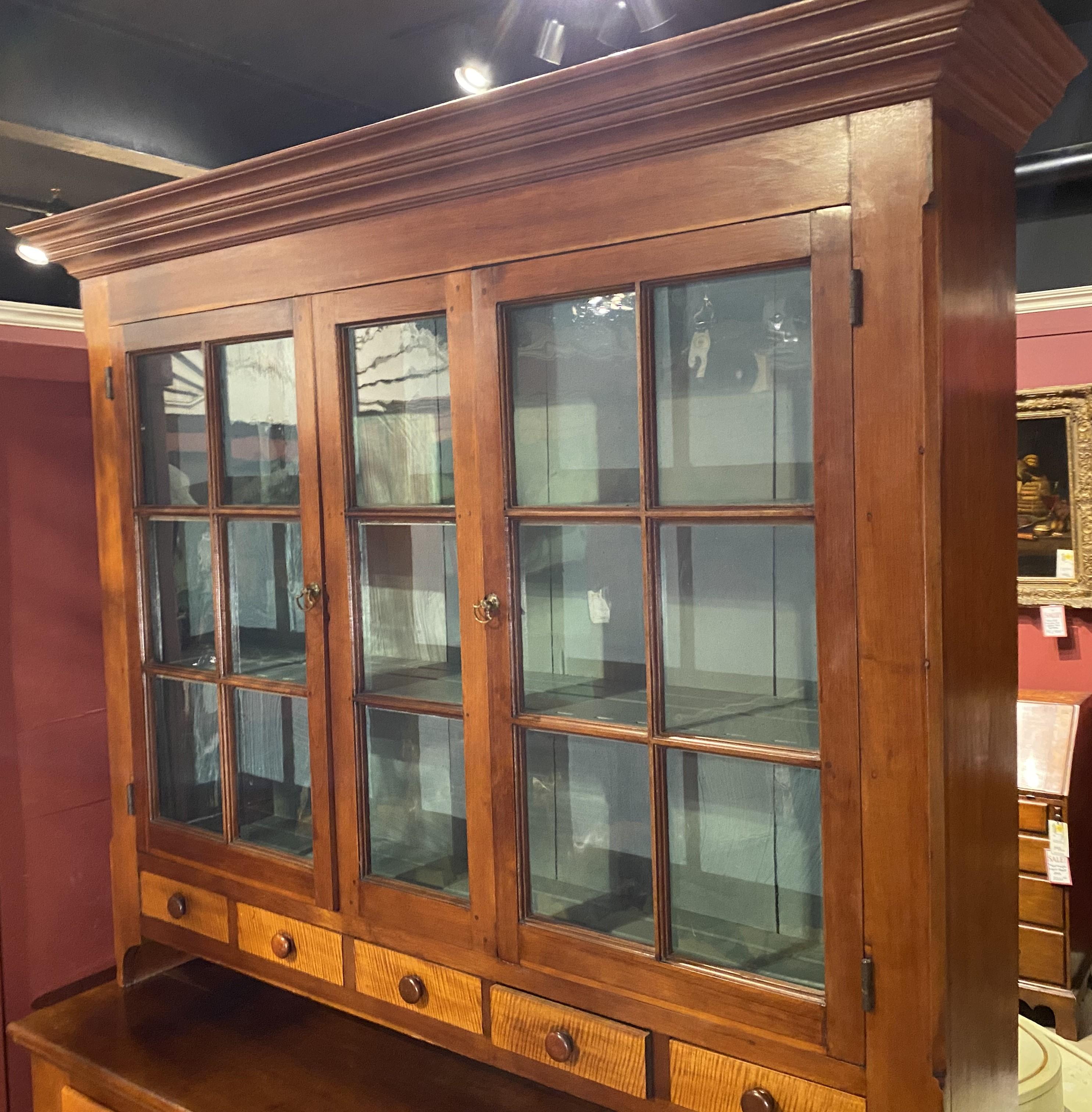 A fine cherry two part step back cupboard, its upper case with a molded cornice surmounting a center three panel window, flanked by two glazed glass doors which open to a light blue painted two shelf interior with carved plate rails, over five small