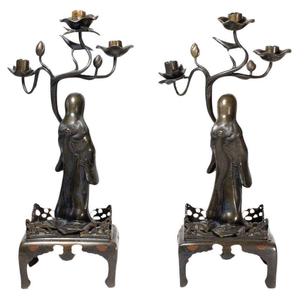 JAPAN - MEIJI period (1868 - 1912) Pair of bronze candlesticks with brown patina with copper and gilded bronze inlays depicting two standing women carrying lotus branches forming a candlestick, posing on quadripod bases decorated with lotus scrolls.