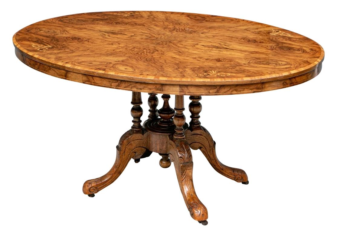 With a book matched burled top, inlaid scrolled decorated banding, and a burled apron. Raised on four turned supports with a turned finial. Mounted on four carved leafy scrolled oak legs on casters. With a protective glass top. 

Dimensions: L. 53