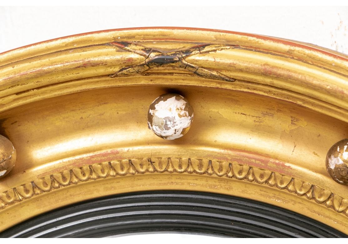 Originally purchased in Boston. A classic deep convex mirror with multiple surrounds. The outer decorated with X shaped motifs on each side, a deep curved band with gilt spheres, an inner thin leafy band, and innermost ribbed ebonized surround.