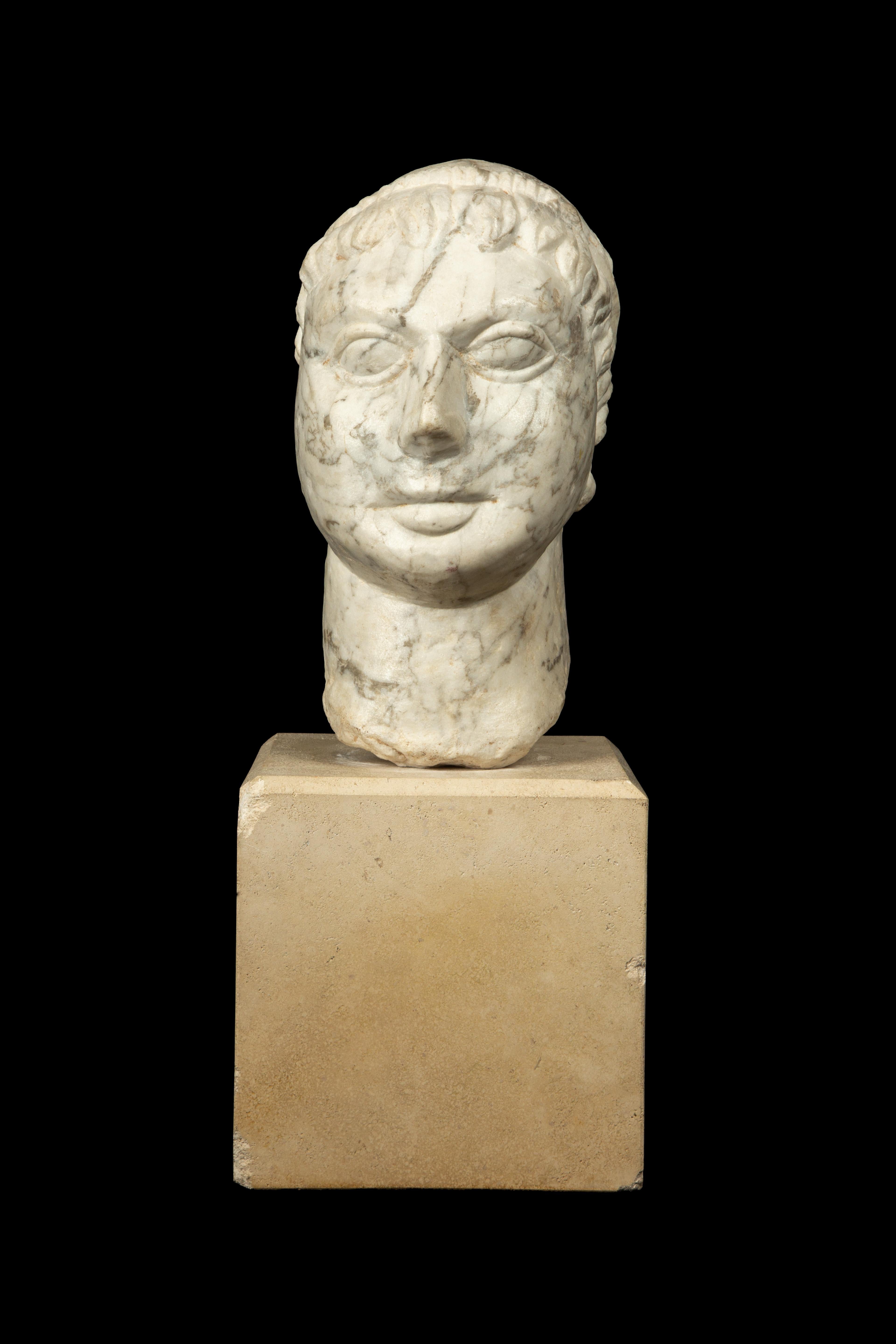 This marble head showcases a man in the antique style, wearing a laurel crown. It is presented on a square stone base, emphasizing its classical elegance and symbolizing achievement. The sculpture is a beautiful reminder of the timeless appeal of