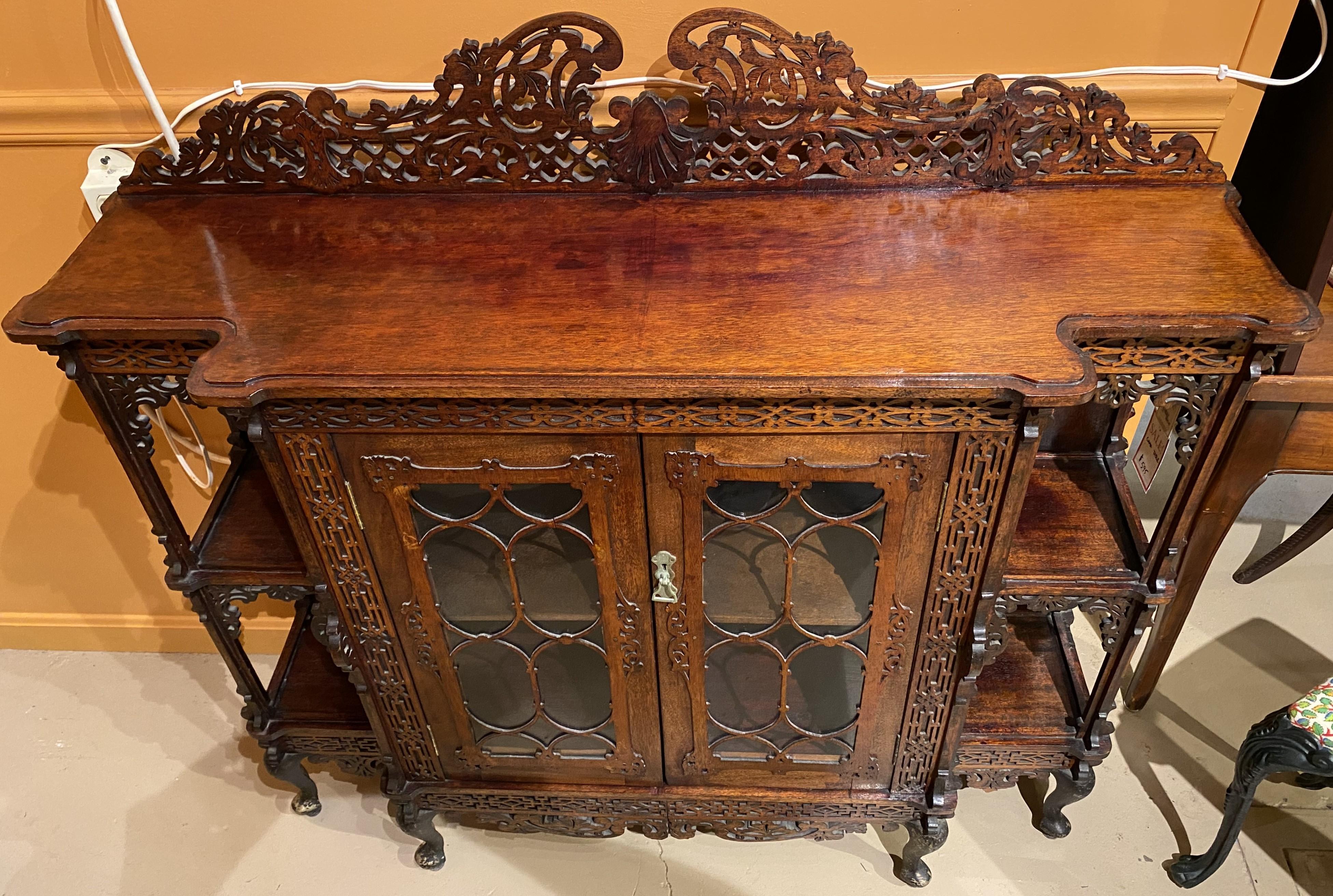 A heavily carved Chinese Chippendale style cabinet étagère with pierce carved scrollwork crest surmounting a conforming case with two glass doors, opening to a single shelf interior, flanked by open shelves on either side, and scroll carved skirt,