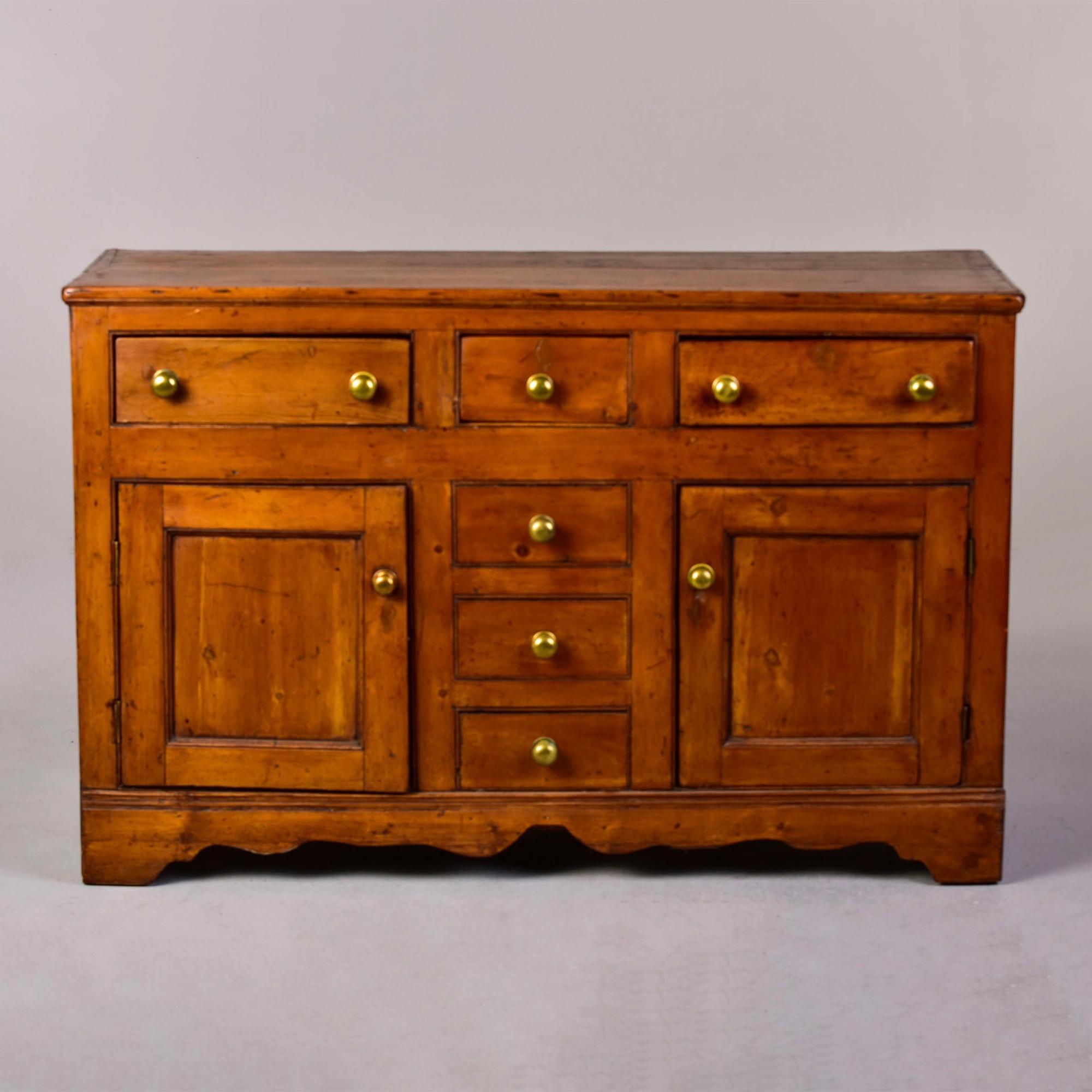 Circa 1880s English country buffet in pine with original brass hardware. Apron at bottom of base has subtle curves. Two lower level hinged door storage compartments with three small drawers between them and the top level has another three drawers.