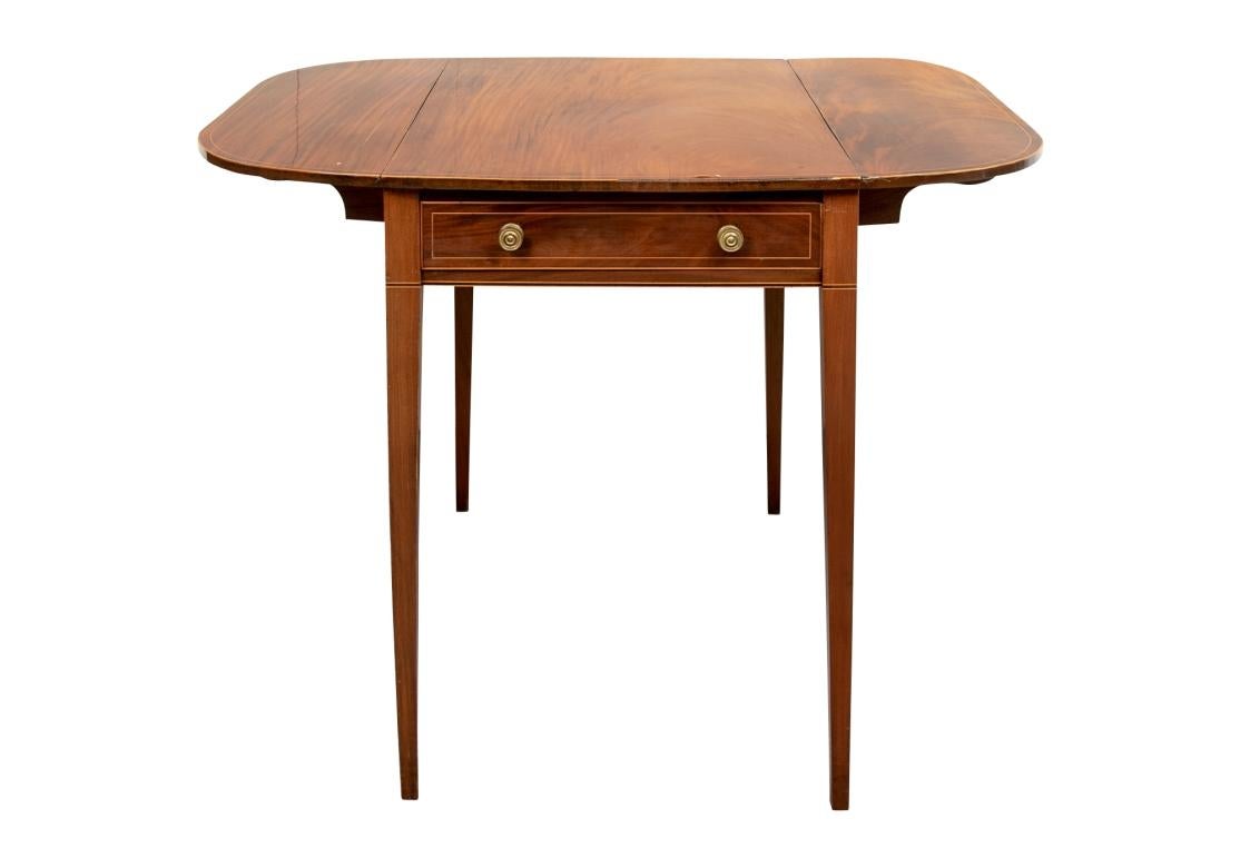 A rectanguar table with slightly curved short ends and rounded drop leaves. The short ends with one working apron drawer and one faux, both with metal knob pulls. Overall fine thin string inlays on the top, drawers and legs. Raised on square