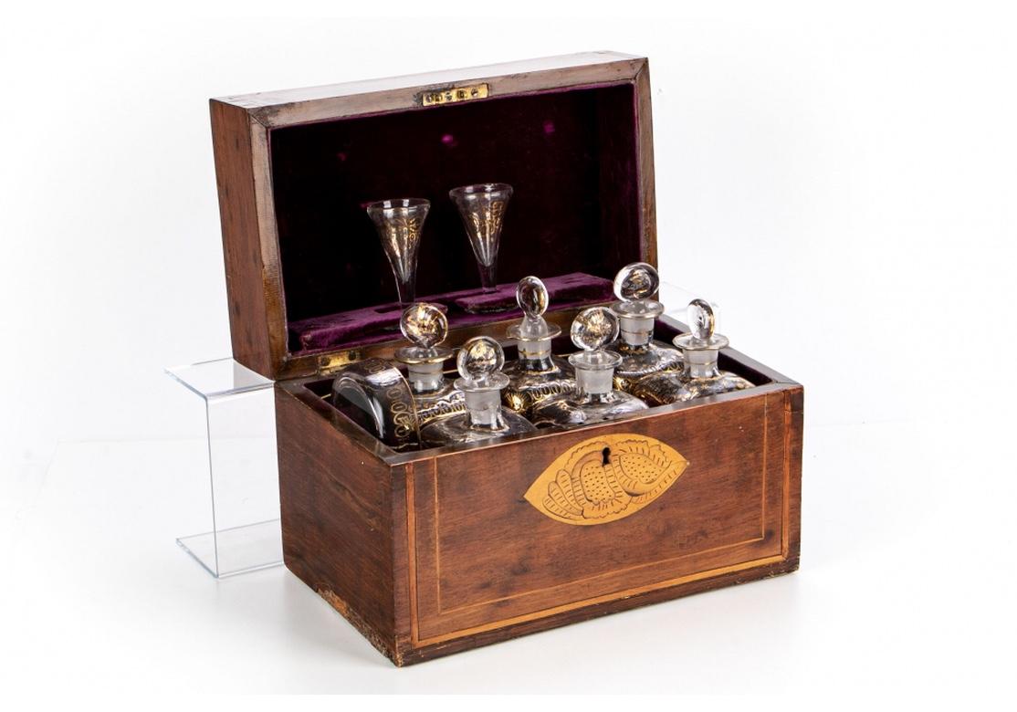 A fine mahogany box with satinwood string inlaid bands and oval incised shell inlays on the lid and front. With six bottles, a bowl, and two small stemmed glasses, all with gilt decoration. With purple velvet lining. 
box L. 12