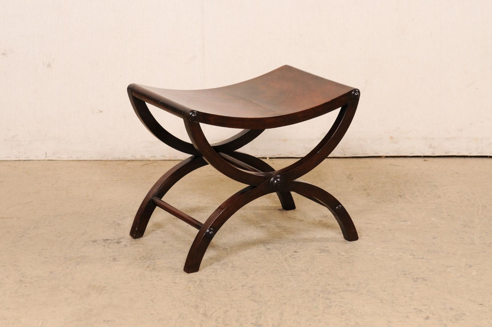An English carved-mahogany wood curule-style stool from the 19th century. This antique stool from England has been created in the Curule (or often referred to as Dante) style, featuring a swag-carved seat which is raised upon a pair of legs