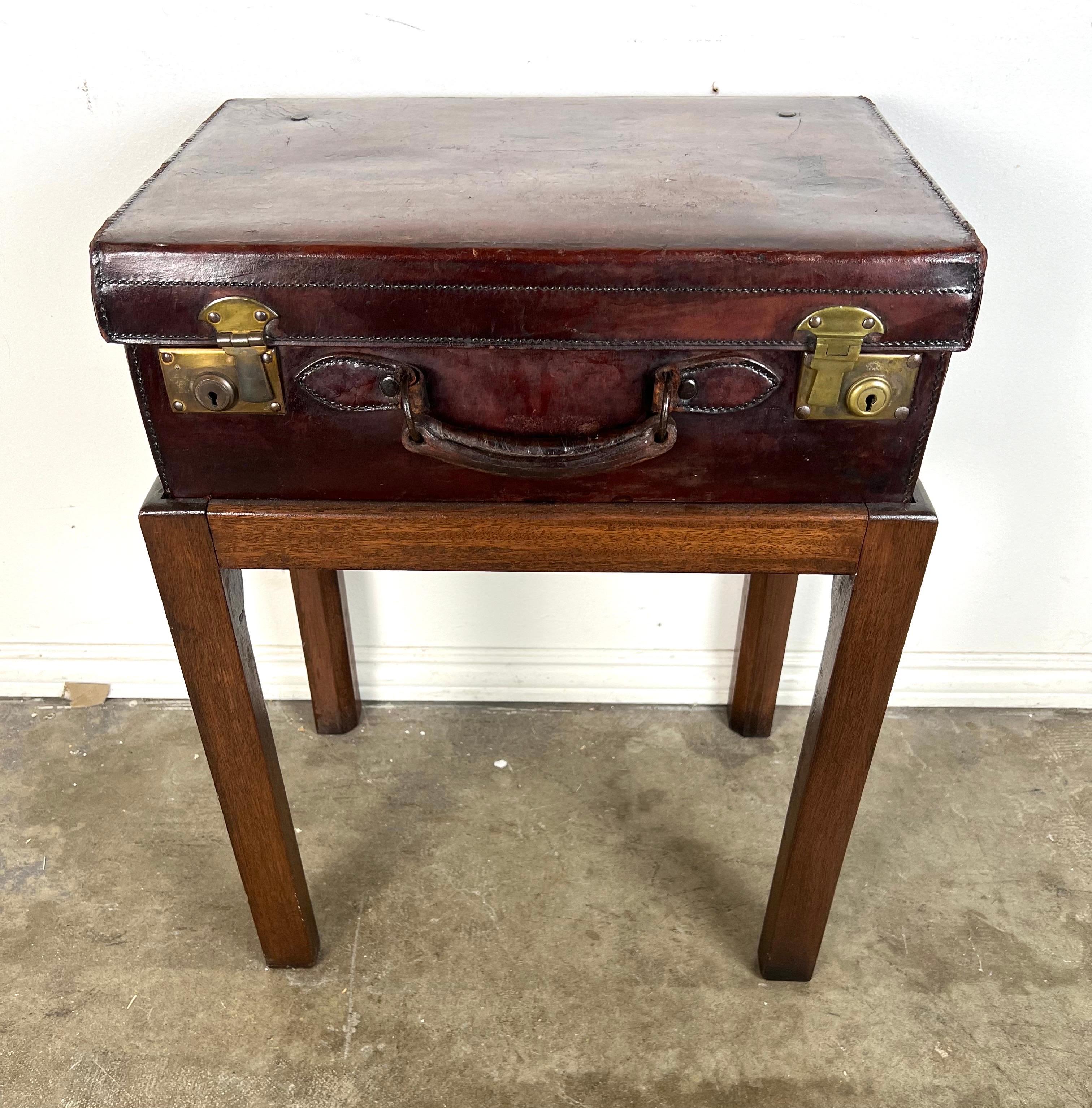 This piece is a unique side table featuring a vintage leather suitcase as the tabletop.  The suitcase has a rich, deep brown exterior with visible wear, giving it a well-loved appearance.  It has brass hardware, including two latches and a central