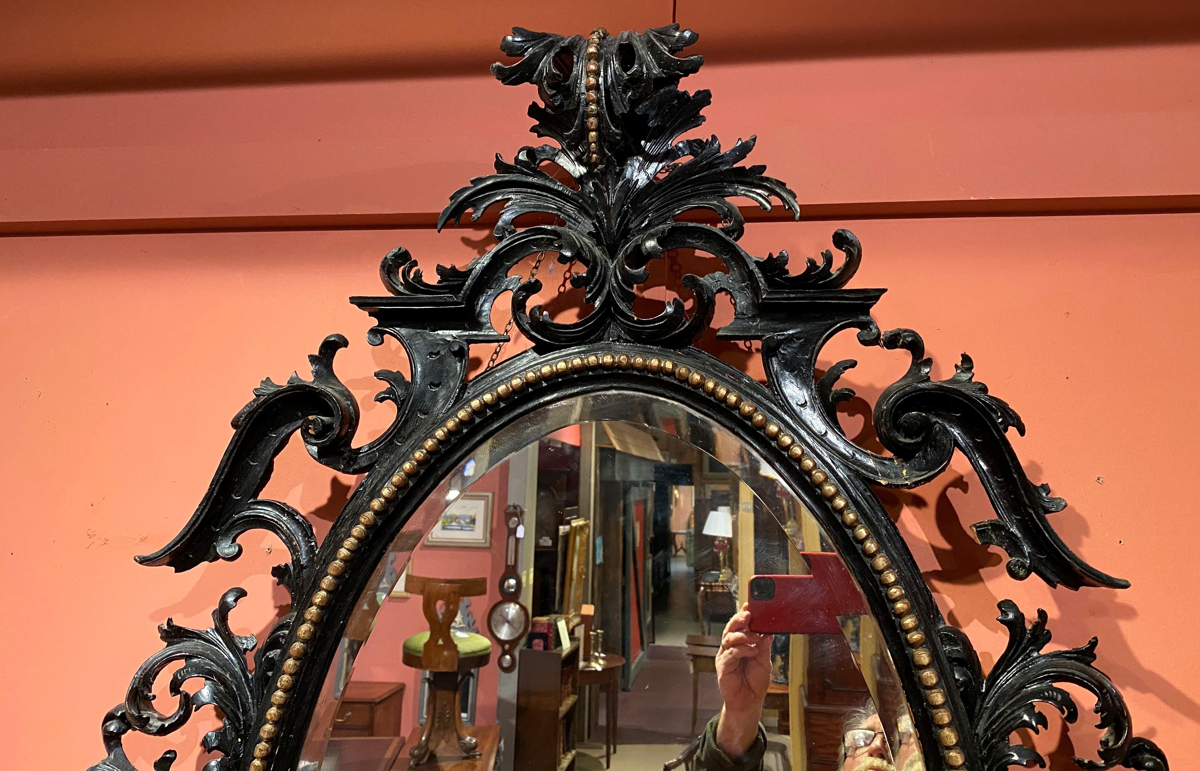A large ebonized oval beveled glass wall mirror in the Chinese Chippendale taste, with heavily carved leaf & scrollwork wooden frame with an overhanging leaf crest and an interior carved gilt sphere border. European in origin, dating to the 19th