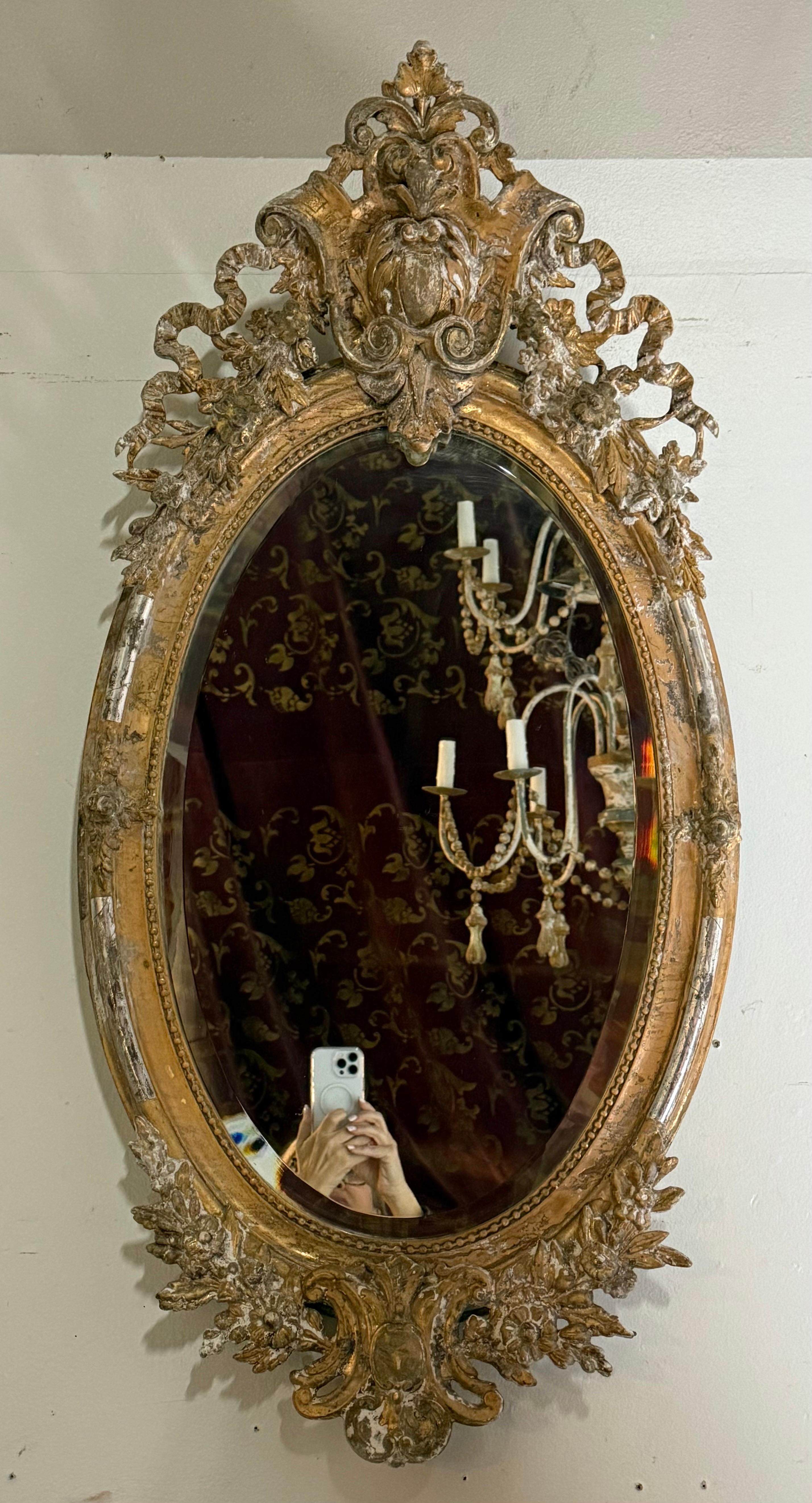 19th century ornate, oval-shaped beveled mirror with highly decorative gilded frame.  The frame features intricate carvings, including floral motifs, scrolls, and a prominent crest at the top with a central cartouche or shield-like design.  The gold