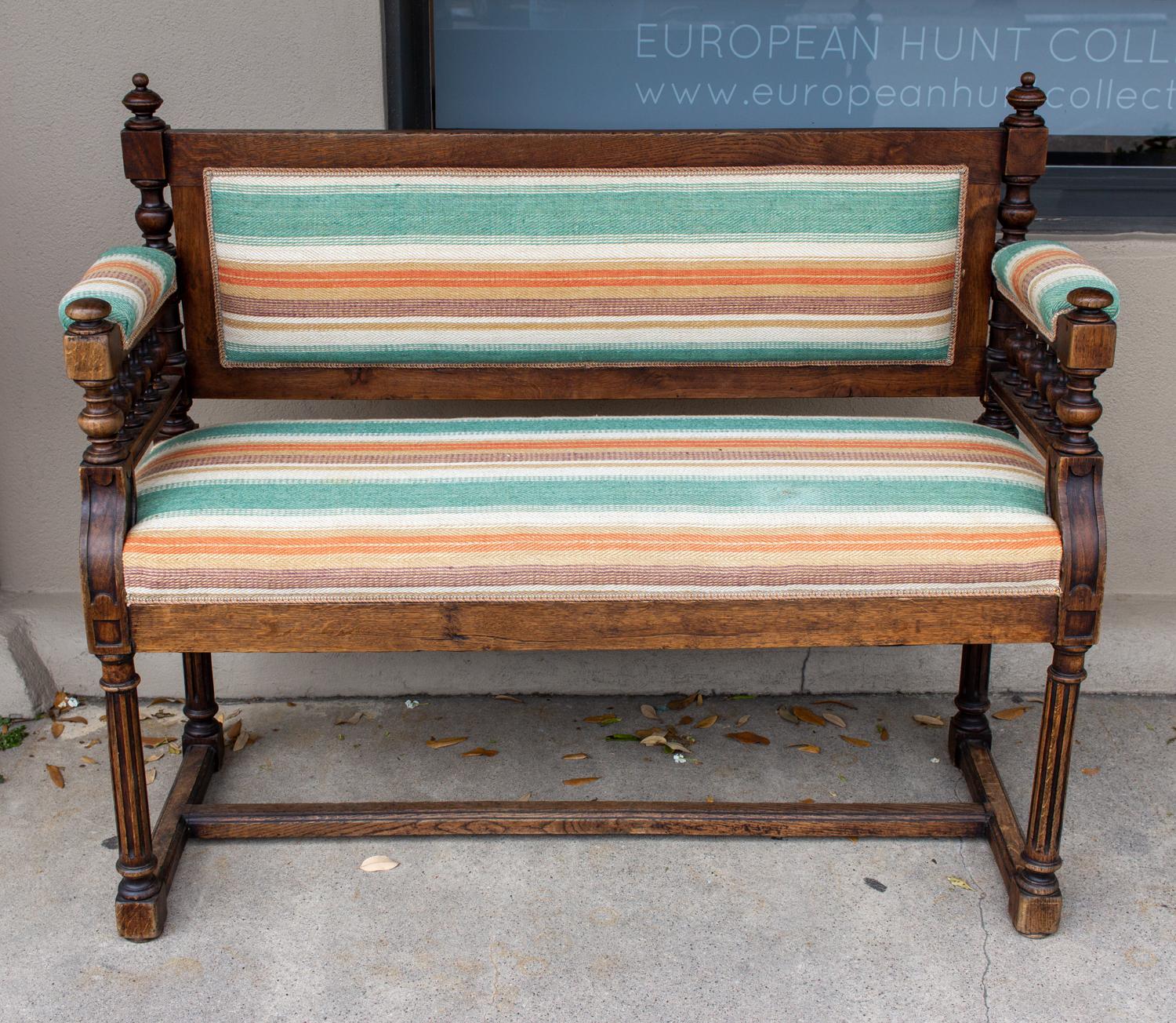 This is an antique French upholstered oak hall bench with striped fabric. The bench has turned spindle details at the arms and finial details on the corners of the back. The upholstery is a striped material in a light orange and spring green stripe