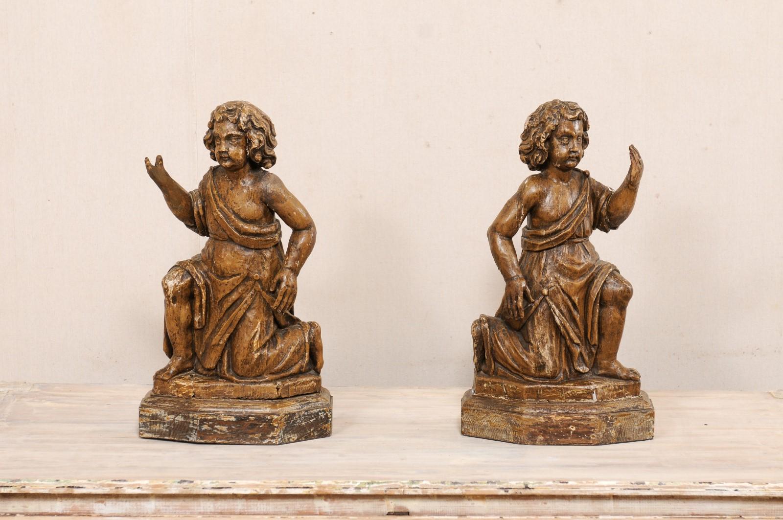 A French pair of carved-wood cherub figures from the 19th century. This antique pair of wooden figurines from France have been hand-carved to depict a male child (or putto) with loosely-curled locks and chubby cheeks, fitted in robed attire, in bent