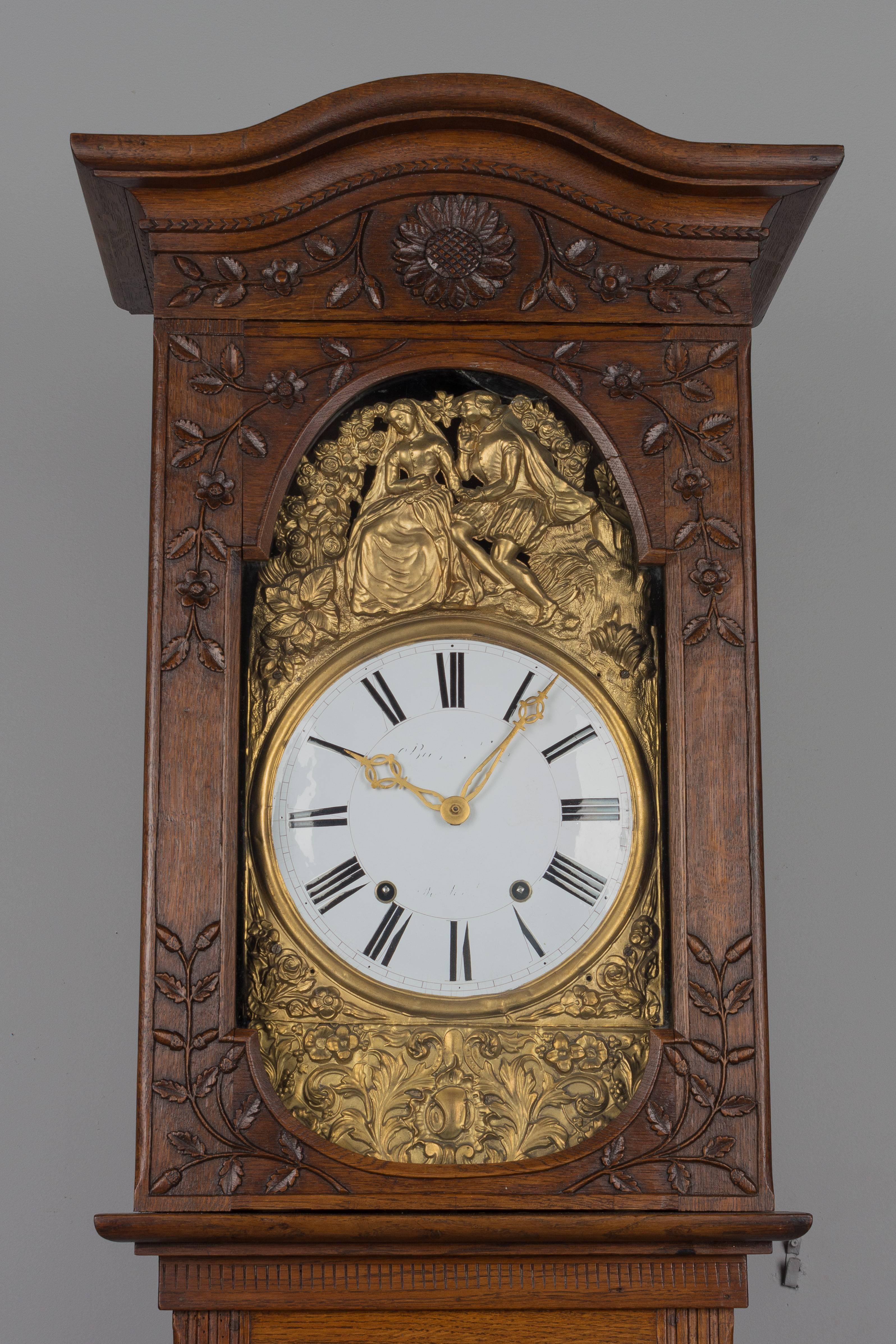 A 19th century French Horloge de Parquet or tall case clock from Normandy. Elegant solid oak case in two parts, with with a chapeau de gendarme crown and fine carvings of oak leaves and acorns. Brass embossment in bright gold has a romantic theme