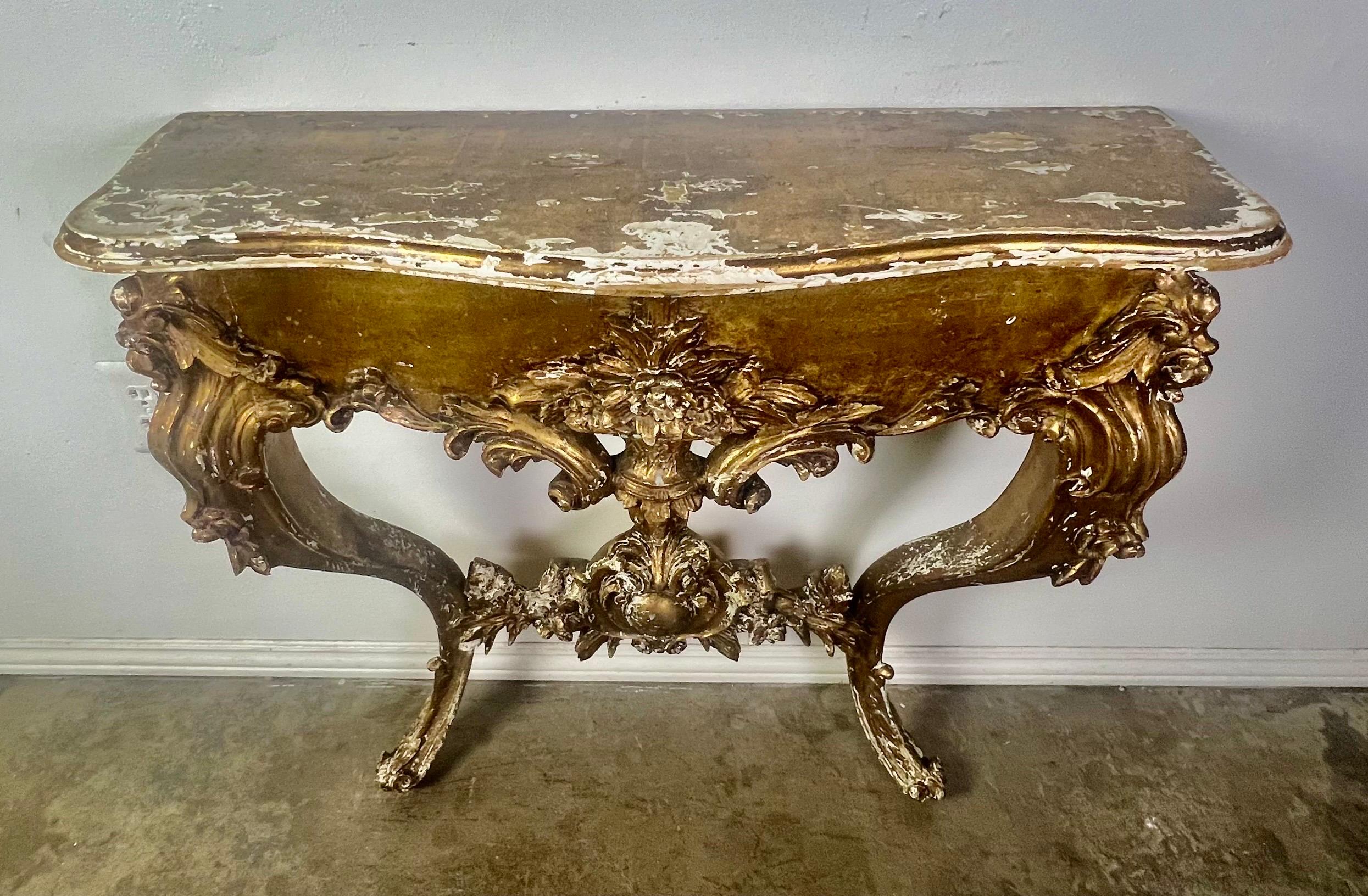19th-century French Louis XV Giltwood console adorned with intricate carvings depicting acanthus leaves, roses, and other detailed elements.  The console stands on cabriole legs that end in rams head feet.  The distressed finish and missing gold
