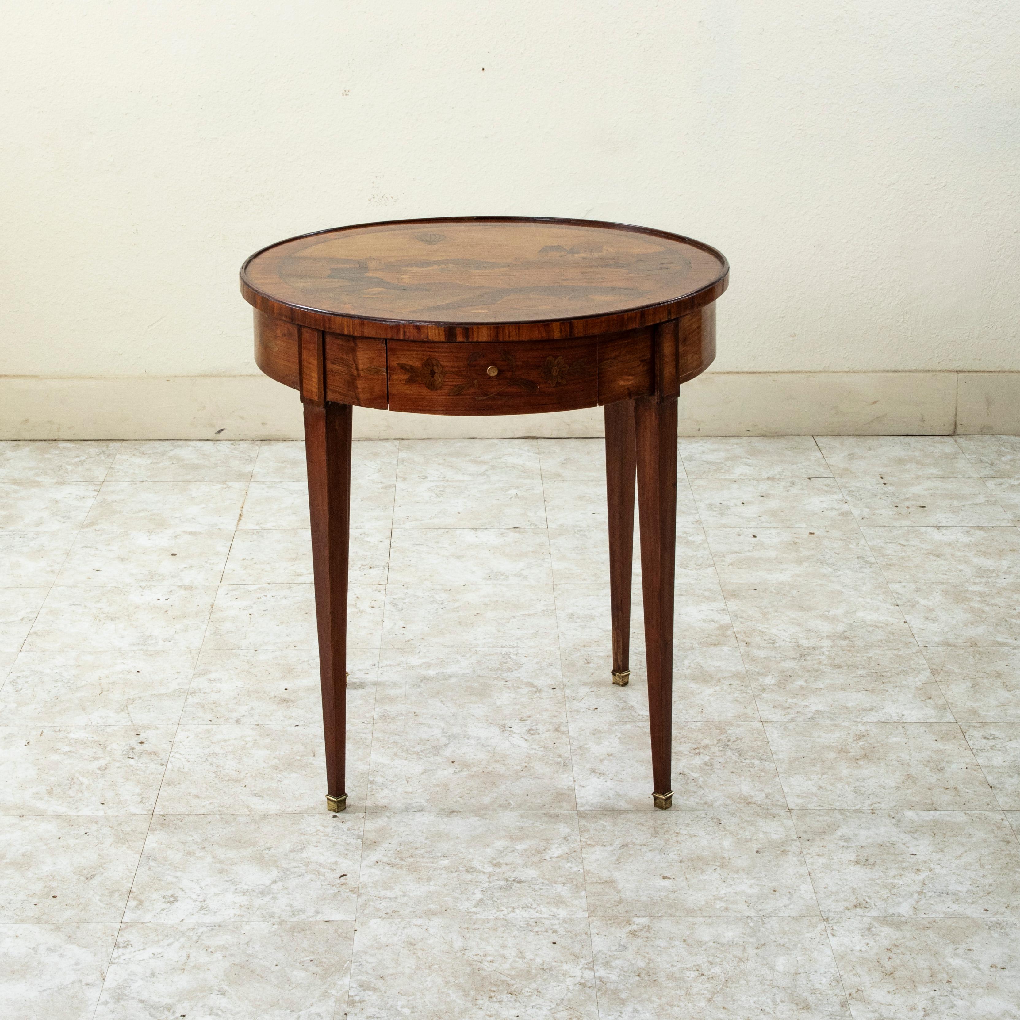 This mid nineteenth century Louis XVI style marquetry side table features a marquetry top in walnut, mahogany, and lemonwood that depicts a genre scene of onlookers at a chateau watching a hot air balloon in flight. The apron displays a a floral