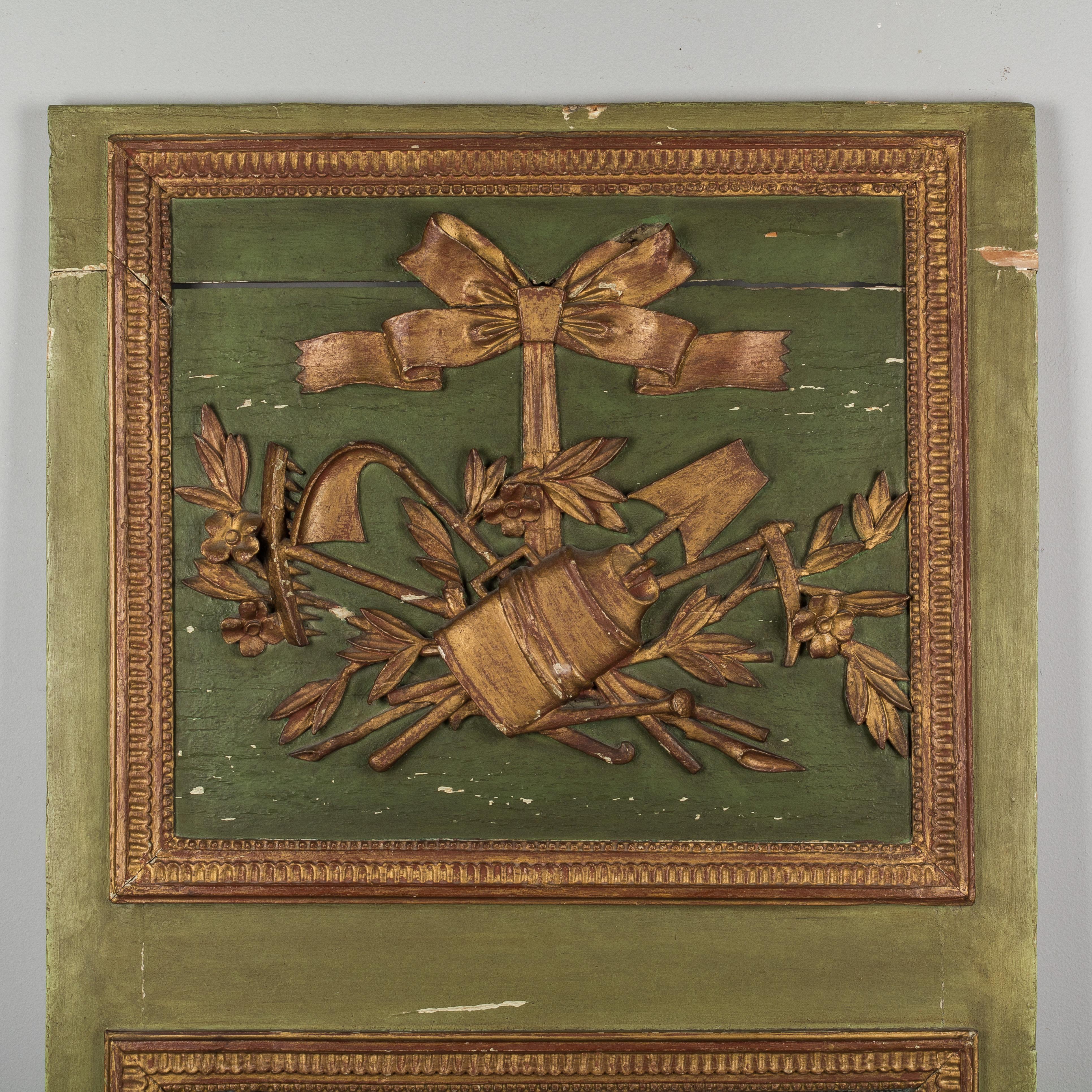 A lovely early 19th century French Louis XVI style trumeau with green parcel gilt frame and carved giltwood relief depicting gardening or farming tools with a large ribbon tied in a bow, flowers, a watering can, rake, and shovels. Perhaps from a sun