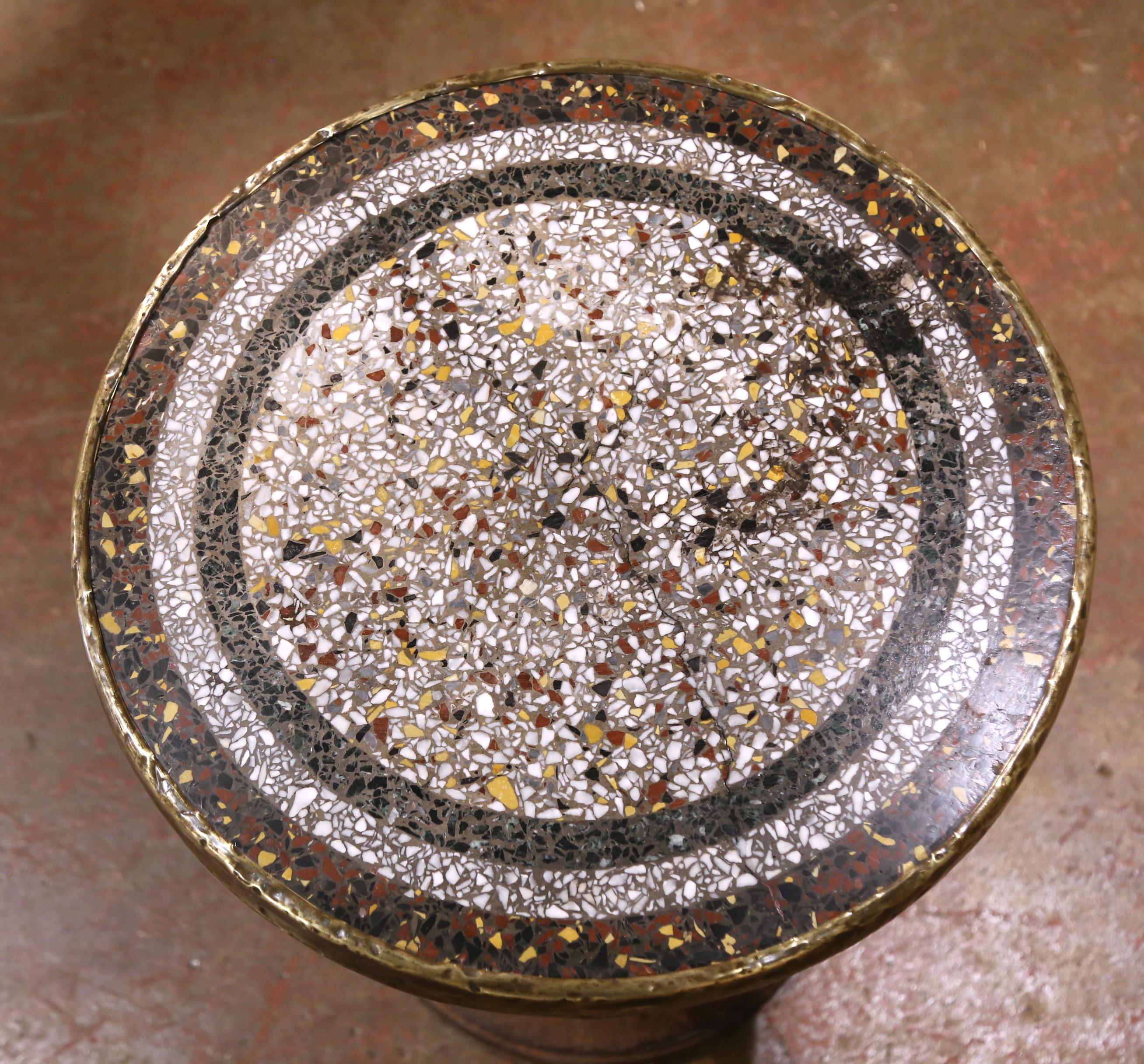 A charming 19th century French bistro table, crafted circa 1870, featuring a colorful terrazzo top inset with marble fragments in tones of white, ochre, black, and red. The circular top is framed with a hand-applied brass band, its gently worn edge