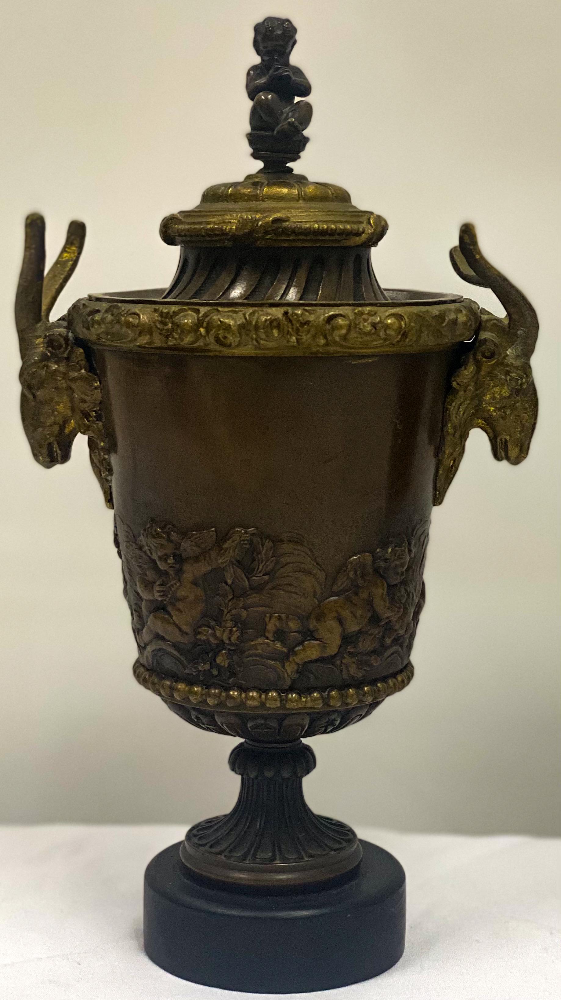 This is a pair of 19th century French neoclassical gilt bronze urns with marble bases. The body of urns are cast bronze encircled with puttis and raised foliate. The handles are gilt bronze ram’s heads, and the finial is a seated putti playing a