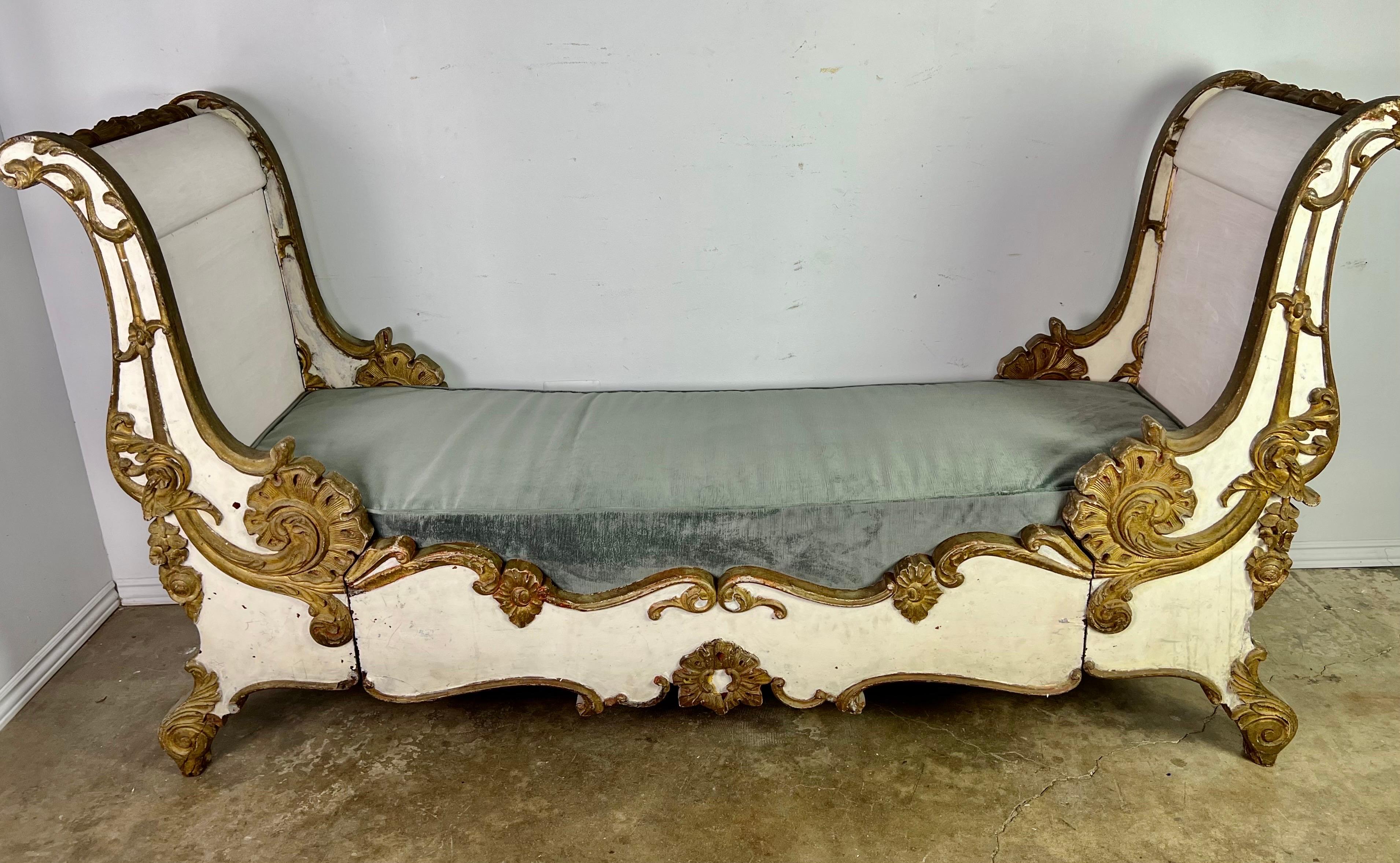 19th C. French Rococo style carved wood daybed. The piece is painted white with gold leaf on the raised carved areas of the bed frame. It is decorated with a floral wreath, acanthus leaf, and floral details throughout. We had a newly upholstered
