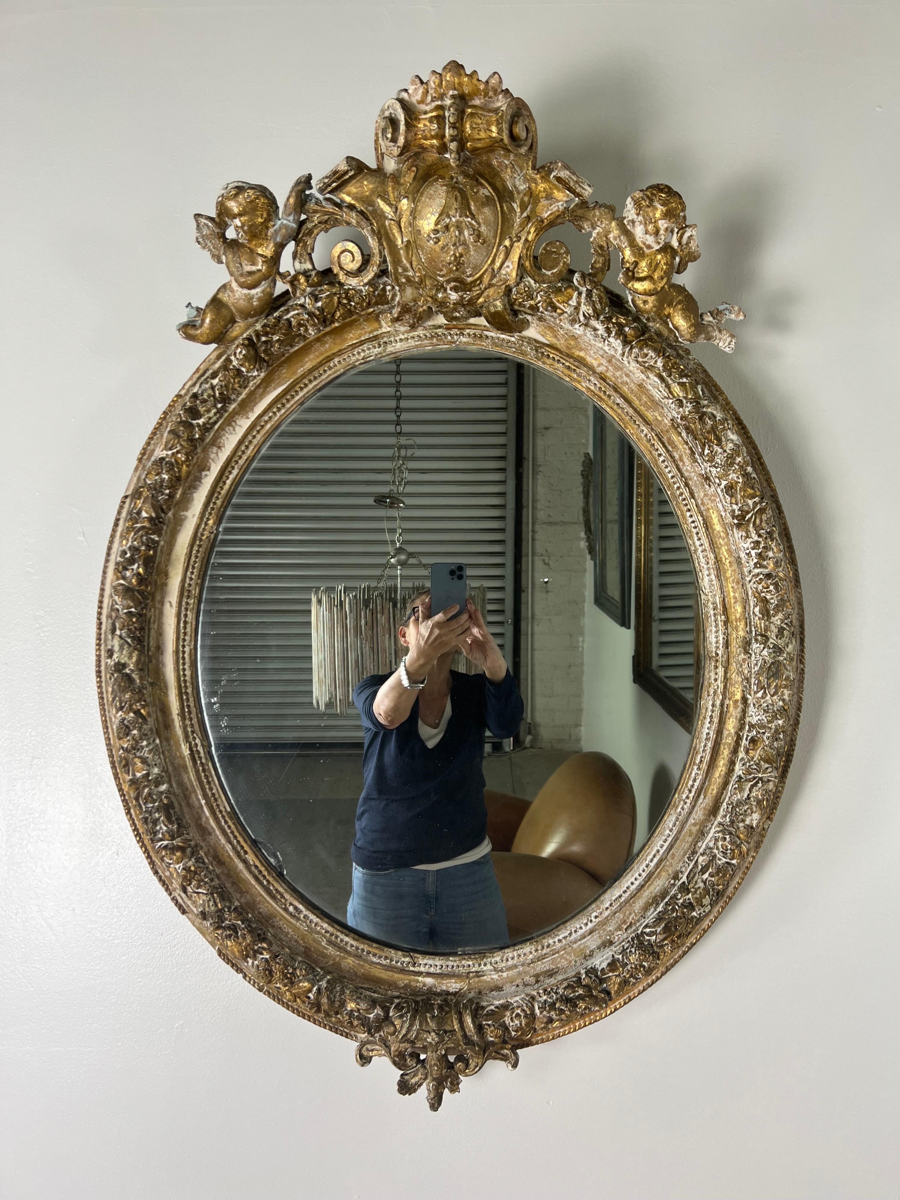 19th century French oval carved wood & gesso mirror with a worn gold leaf finish. There is a cartouche on the top that is flanked by two cherubs. The mirror is original and has begun to antique.