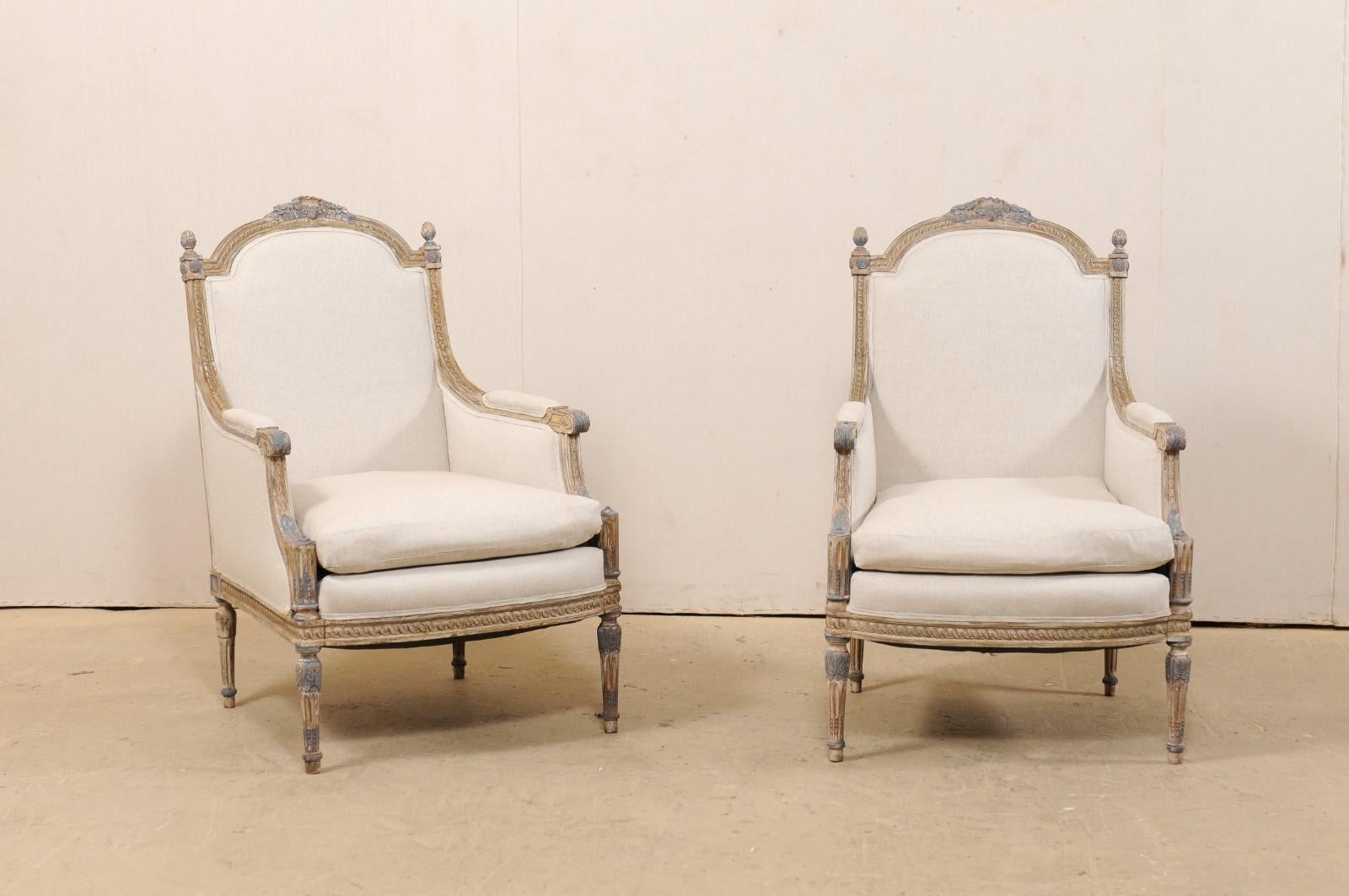 A French pair of occasional bergère chairs from the 19th century, with new upholstery. This antique pair of Louis XVI style armchairs from France each features a beautifully carved arched back-rail with foliage crest at center, a prominent pair of