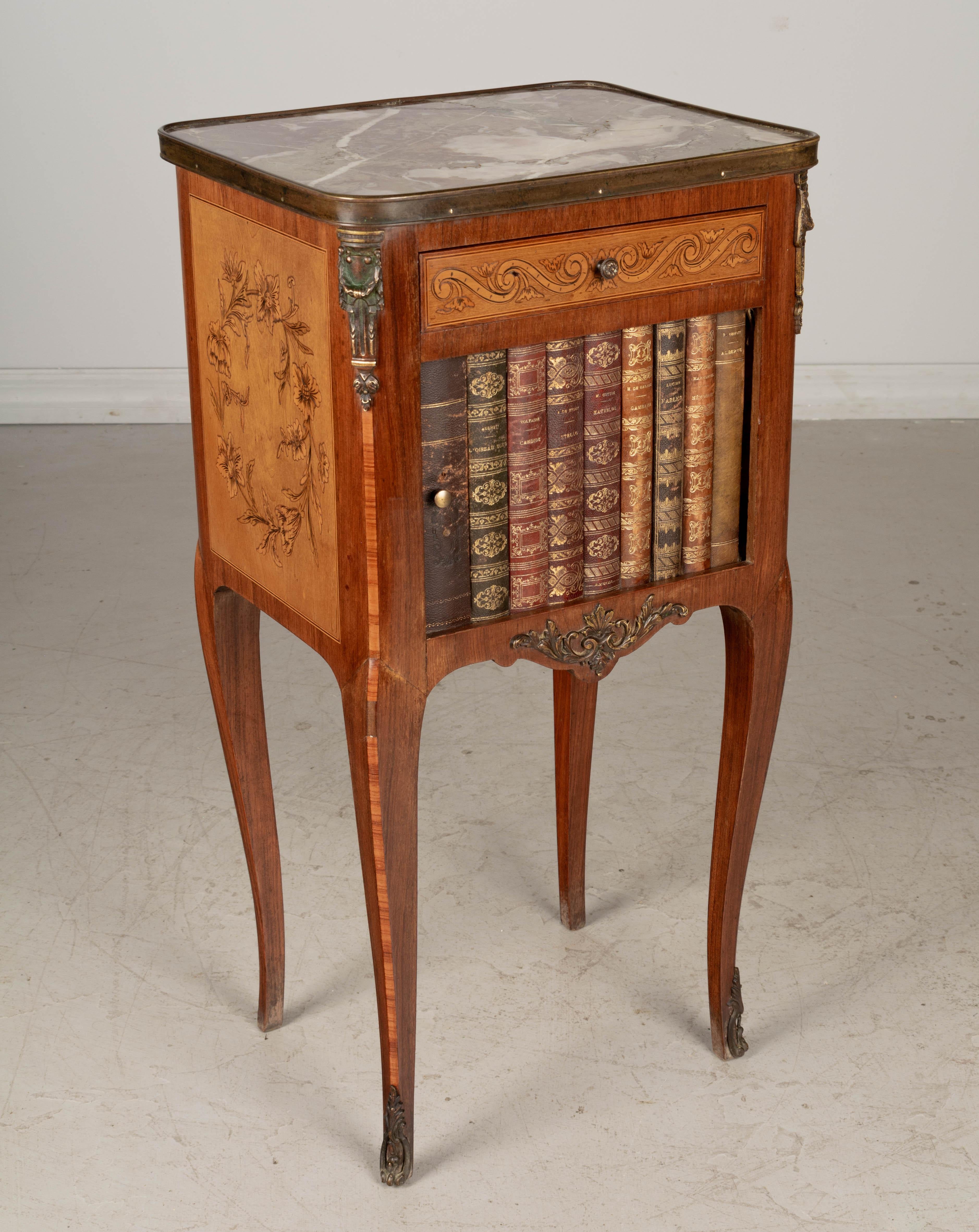 A late 19th century Louis XV style French bronze mounted marquetry bedside table with leather faux livres tambour door, small dovetailed drawer and marble top. The tambour door is faced with gold embossed leather bound book spines with titles by