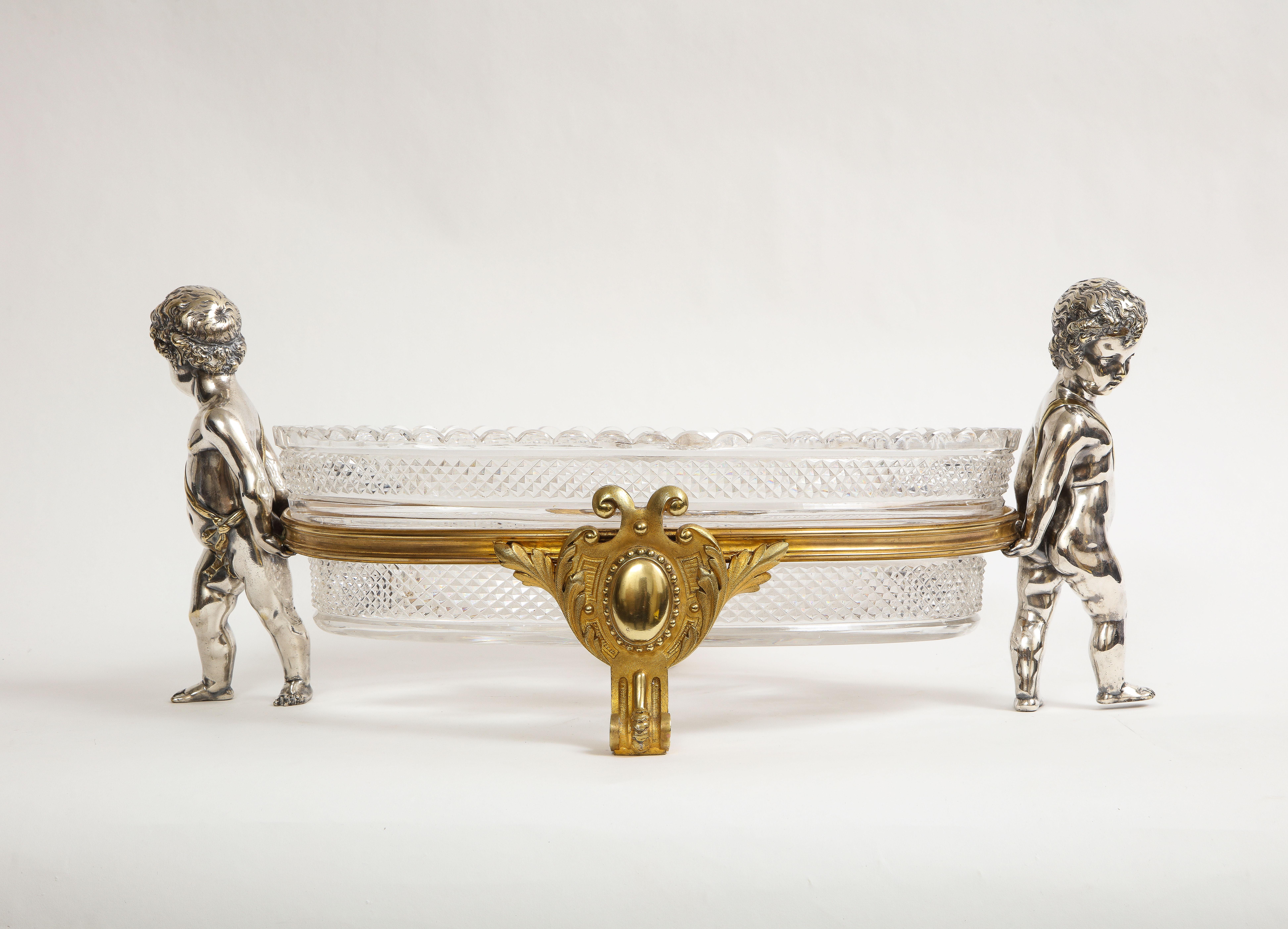 A large Louis XVI Style French Silvered and Gilt Bronze Putti Mounted crystal centerpiece, Marked Baccarat, from the 1800s. The crystal centerpiece is being held up by two silvered bronze putti's which are seen wearing bands around their bodies and