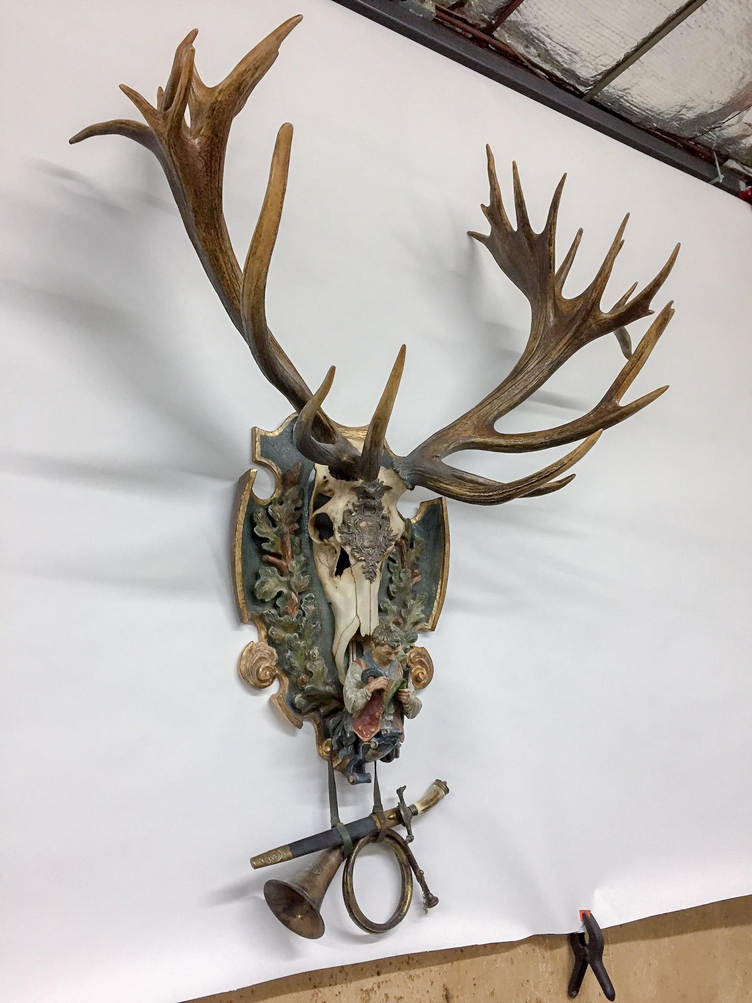This historic, 19th century German red stag trophy is crafted with a hand carved polychrome Black Forest plaque featuring full cap, decorative plate hardware, Fürst Pless hunt horn and Hirschfänger. The carving at the bottom of the plaque represents