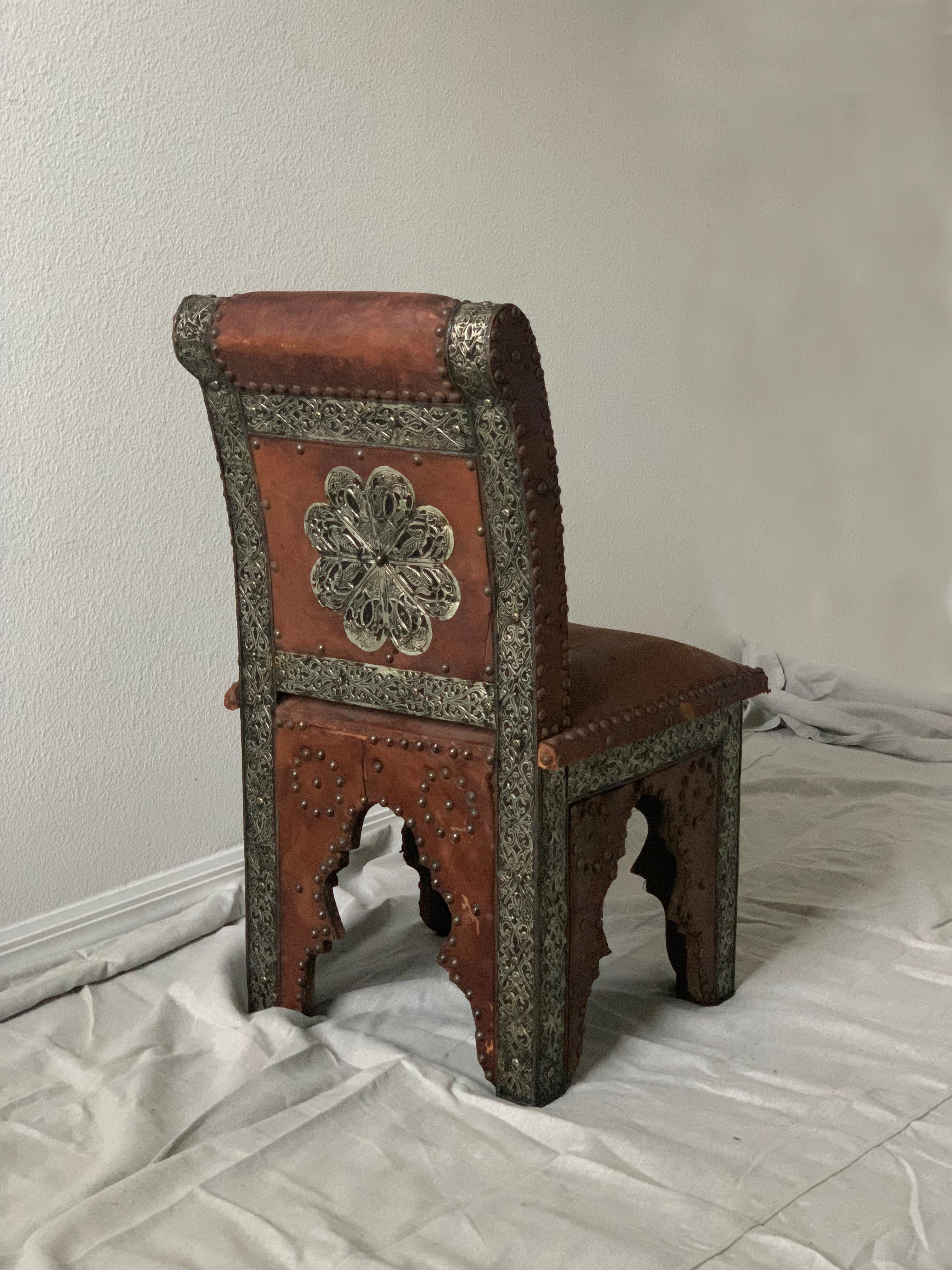 Upholstered in leather, chair displays intricate detailing - hand hammered metal, various studded detailing. Chair frame is wood and is raised on square inlaid legs with carved Arabic arches.