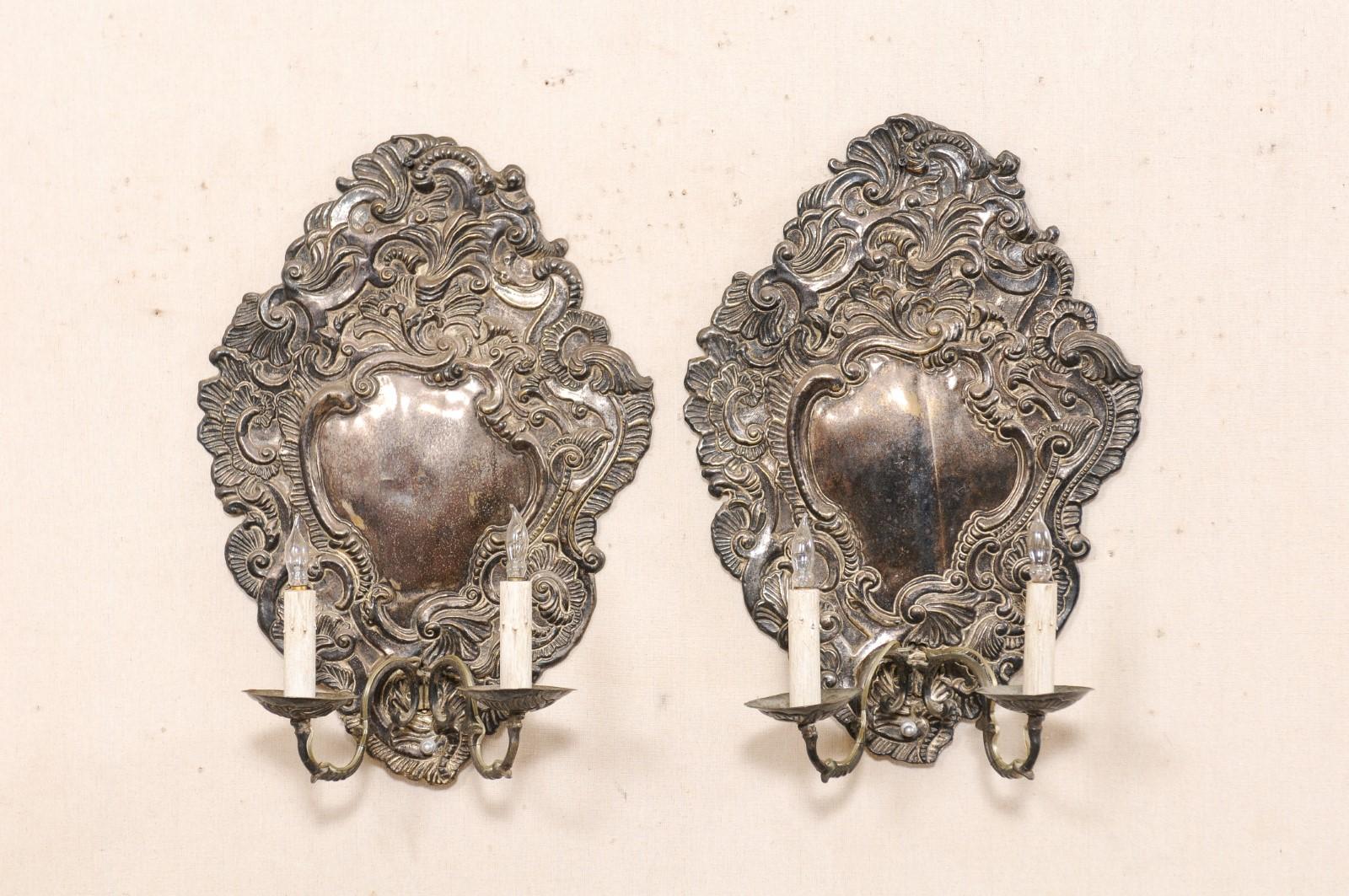 An Italian pair of Baroque style silver plated, two-light wall sconces from the 19th century. These antique wall lights from Italy each feature shapely back-plates in the elaborate Baroque design and swirling movement of stylized shells and foliate
