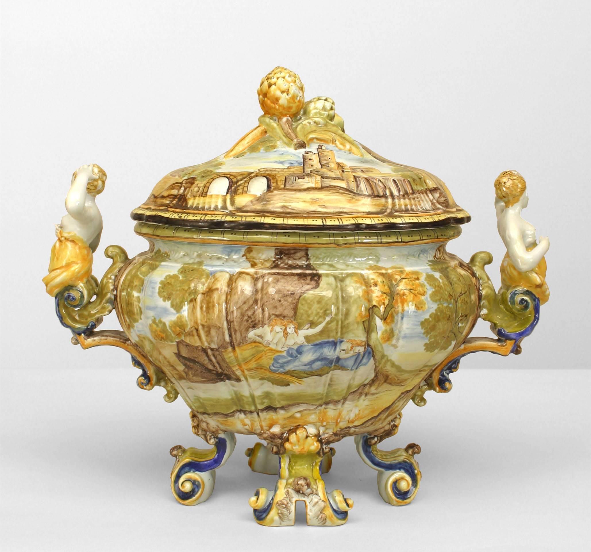 Italian (19th Century) Majolica porcelain tureen decorated with a landscape of figures and castle with female figural handles and supported on 4 scroll feet with a fruit finial top.
